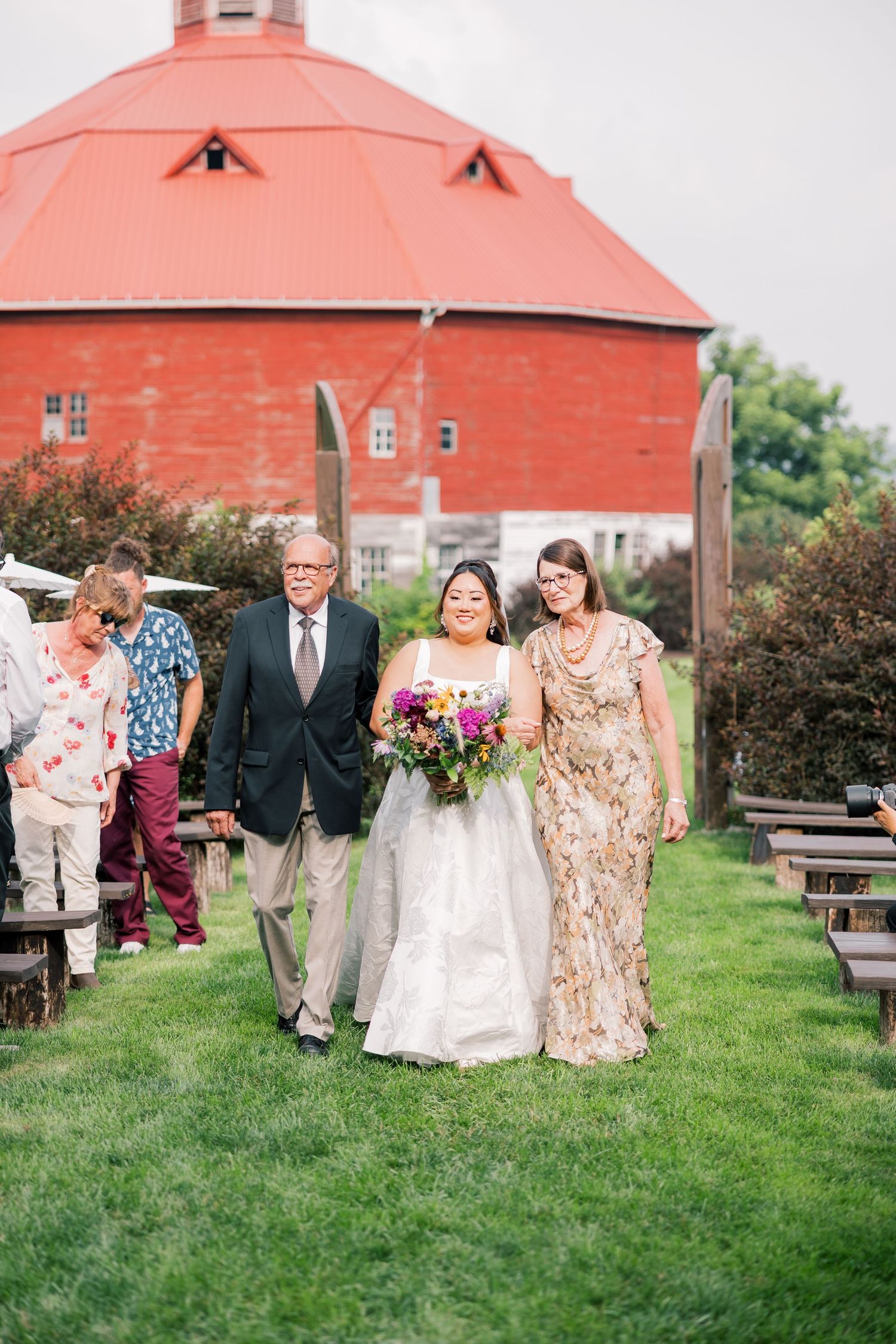 Wedding party poses for photos outside a large red barn on a grassy lawn.
