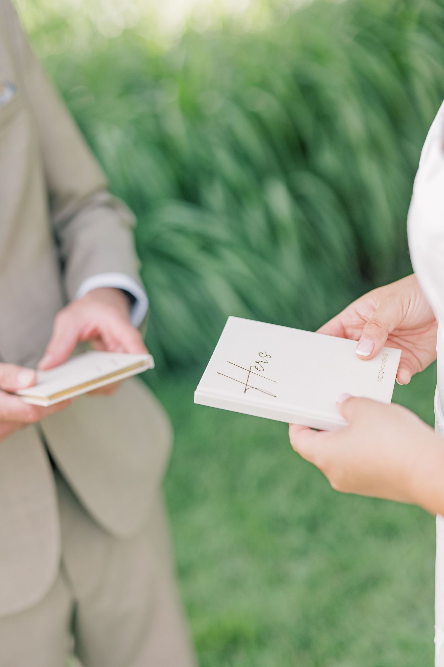 Close-up of hands exchanging wedding vows written in small white books outdoors.