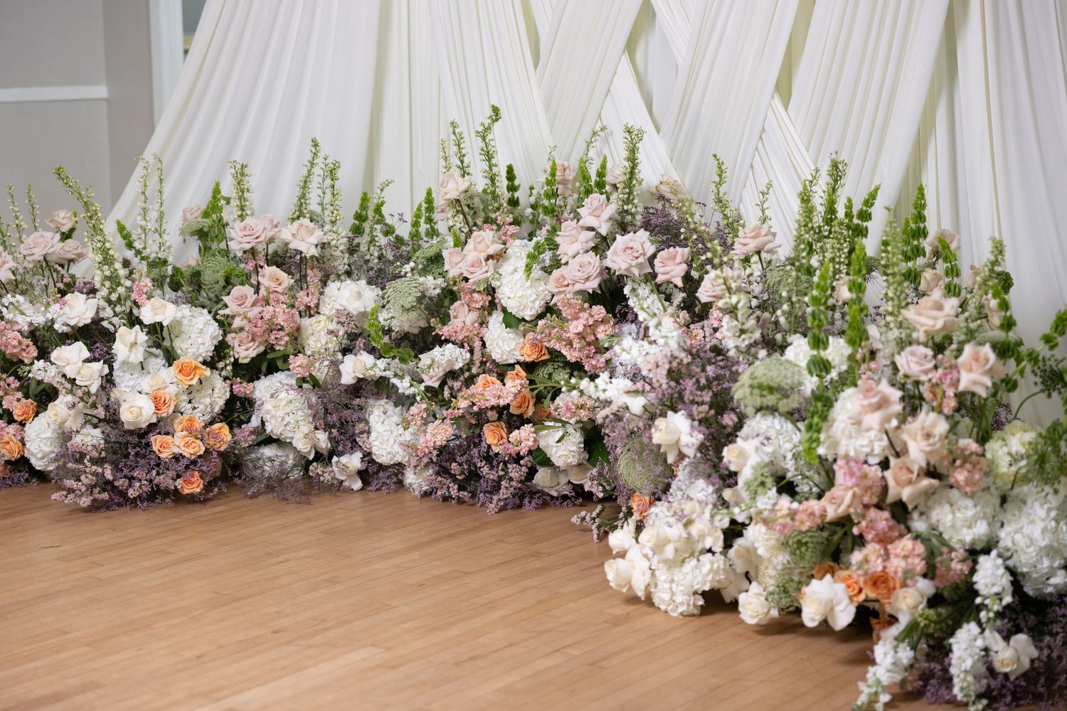 Elegant floral installation with white and blush roses cascading along a white wall and floor for a wedding ceremony.