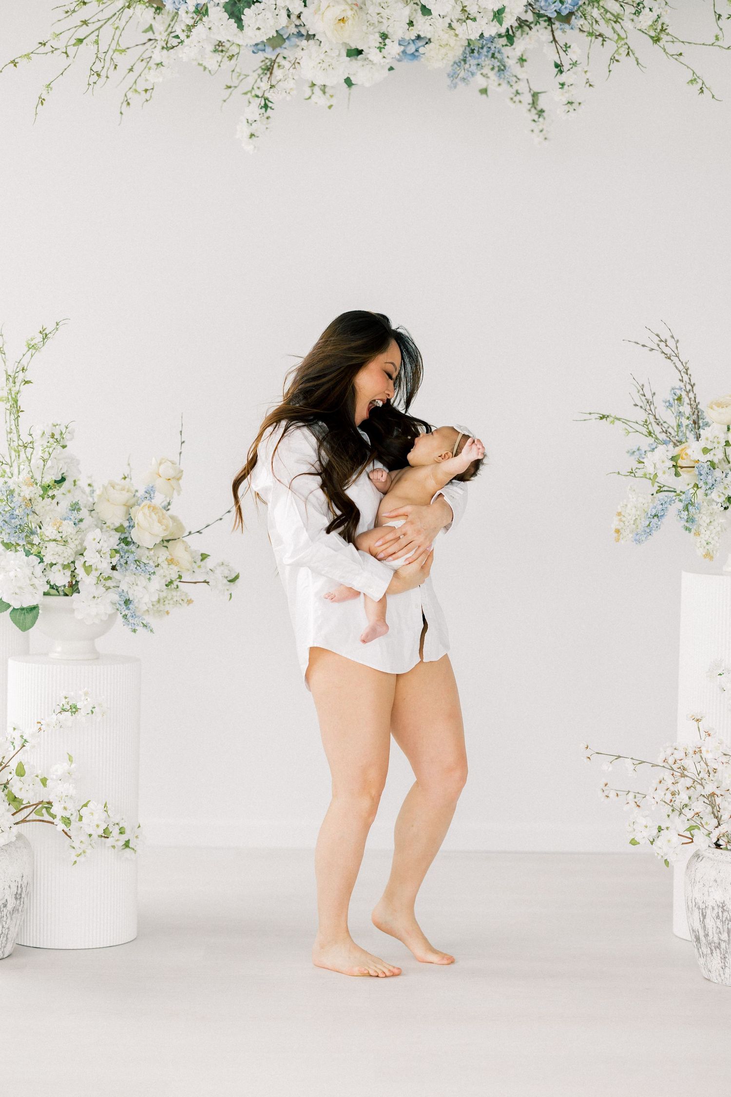 A mother in a white shirt kisses her newborn baby surrounded by elegant blue and white floral arrangements.