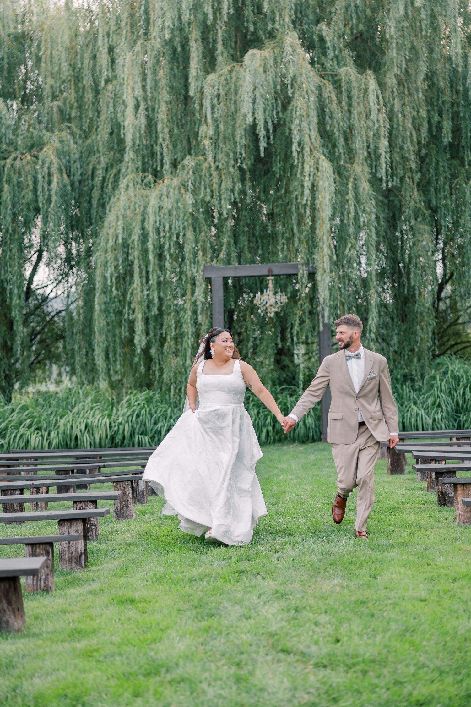 A couple walks hand in hand through a garden ceremony site with weeping willow trees.