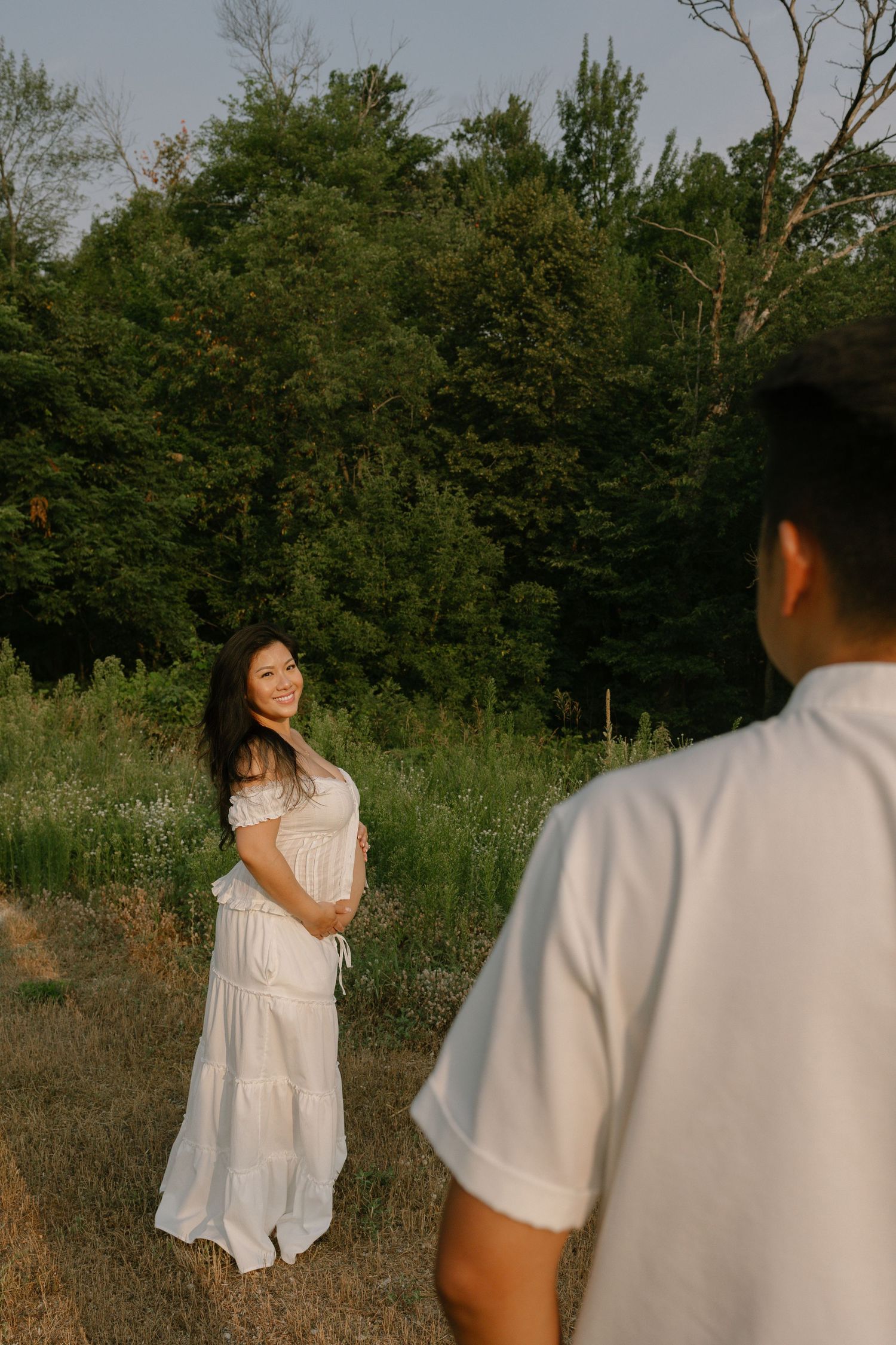 A smiling pregnant woman in a white dress stands in a sunlit meadow as her partner approaches her.