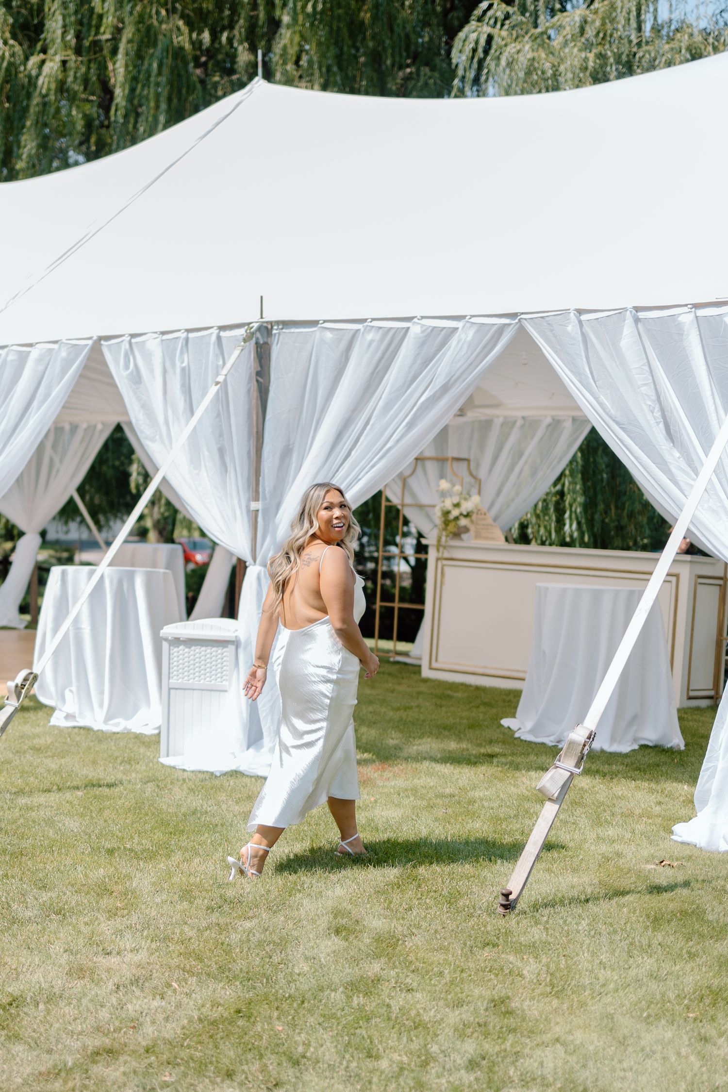 Playful wedding party members peek through flowing white tent drapes on a sunny lawn with willow trees in background.
