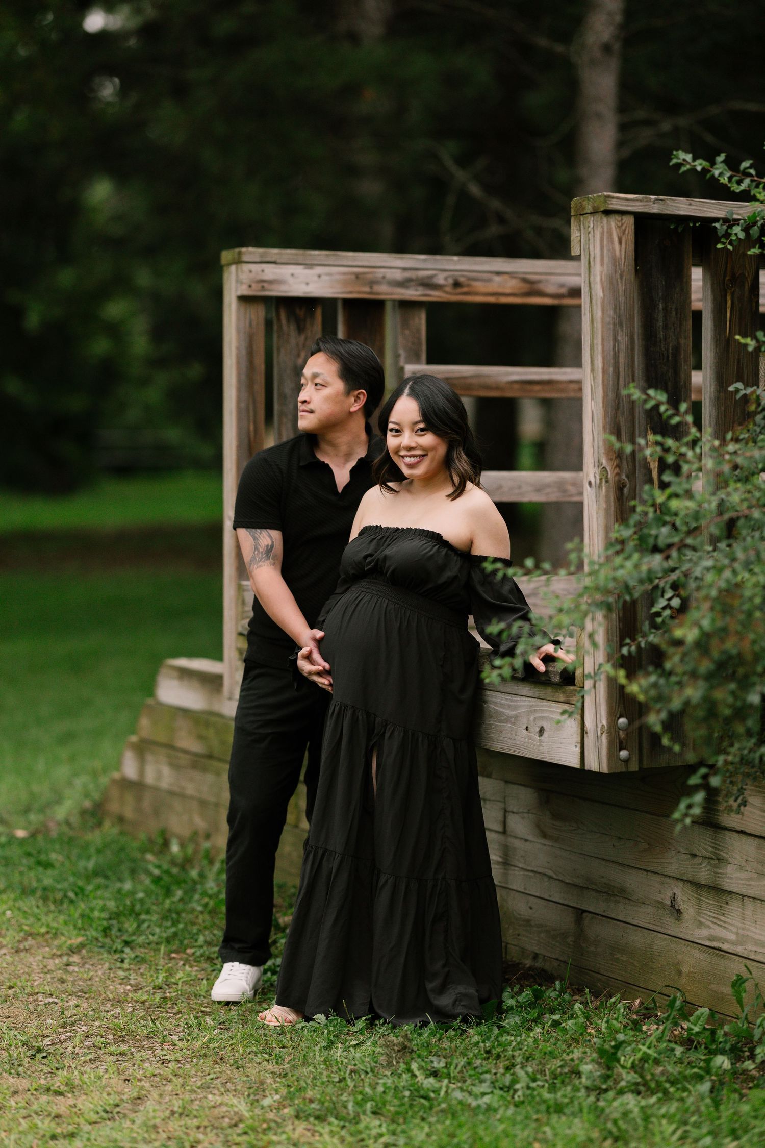 Two women in black dresses pose together on a rustic wooden fence in a lush garden setting.