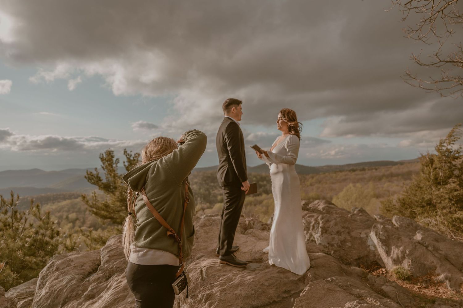 A photographer captures a wedding ceremony on a dramatic mountain overlook with stormy clouds in the background.