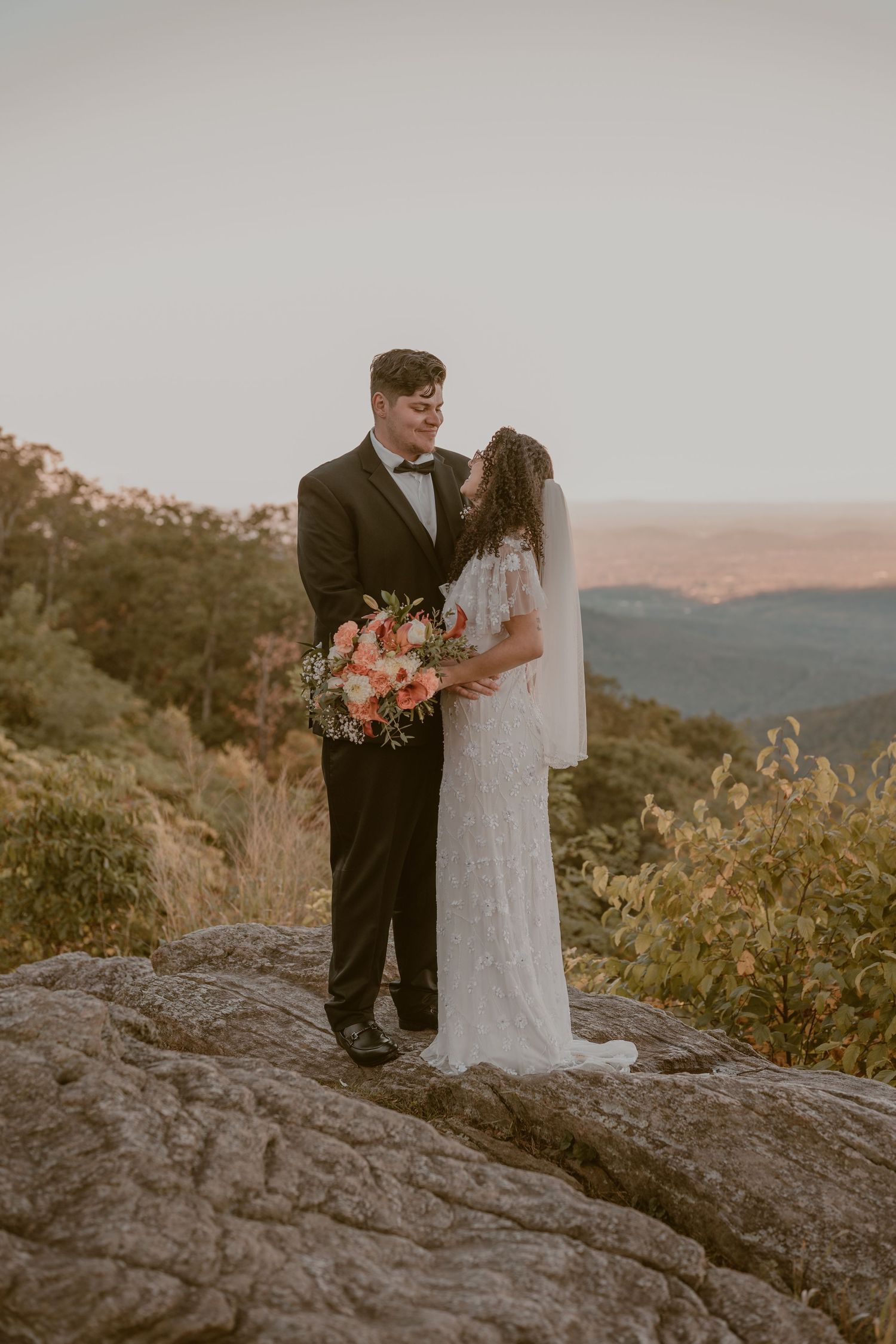 A couple in wedding attire stands on a rocky mountain overlook at sunset with scenic mountain views in the background.
