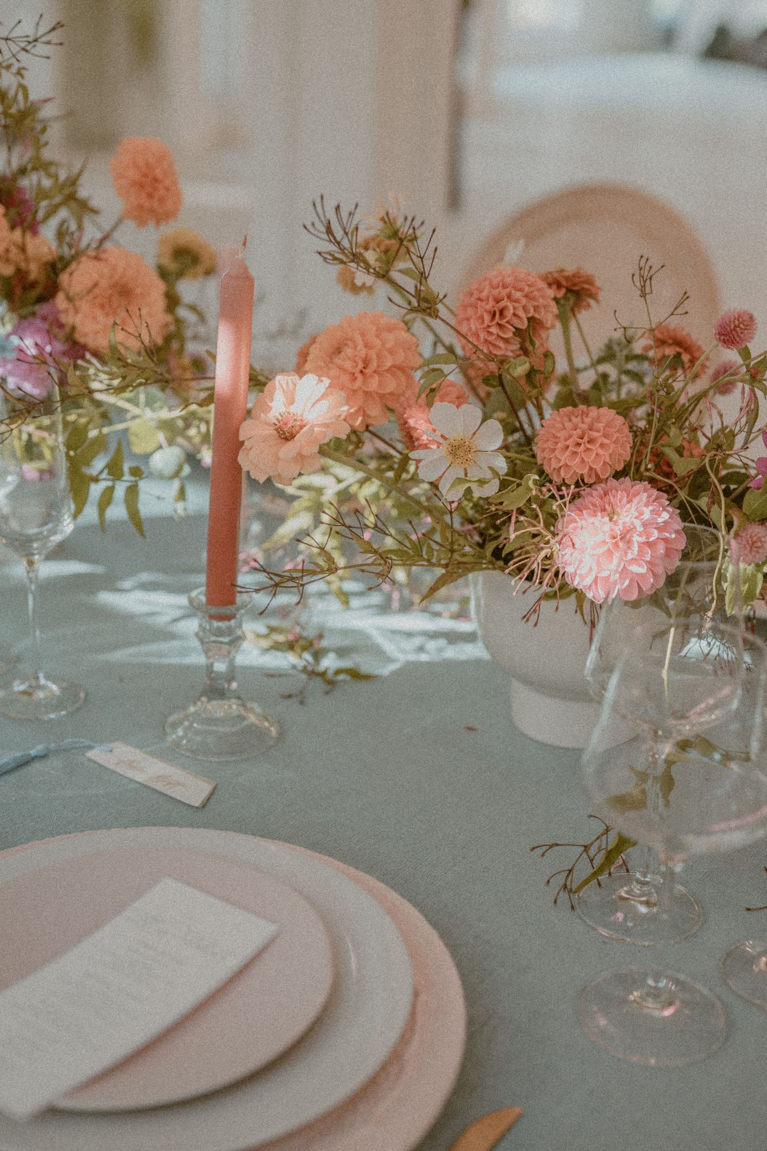 Elegant table setting with coral pink carnations and delicate glassware on a pale gray tablecloth with soft candlelight.