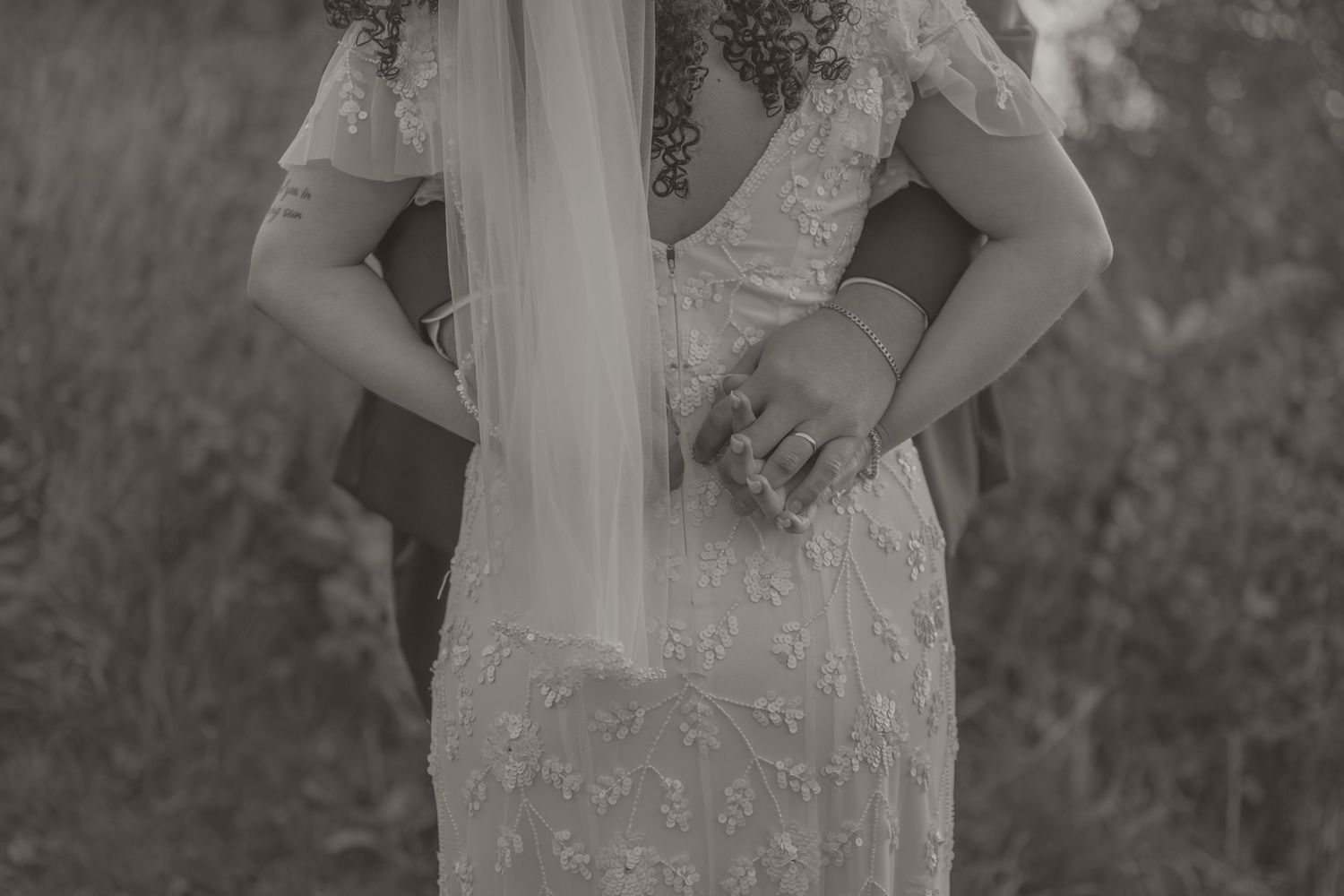 Black and white detail of a vintage-style lace wedding dress with delicate beading and flowing veil in an outdoor setting.