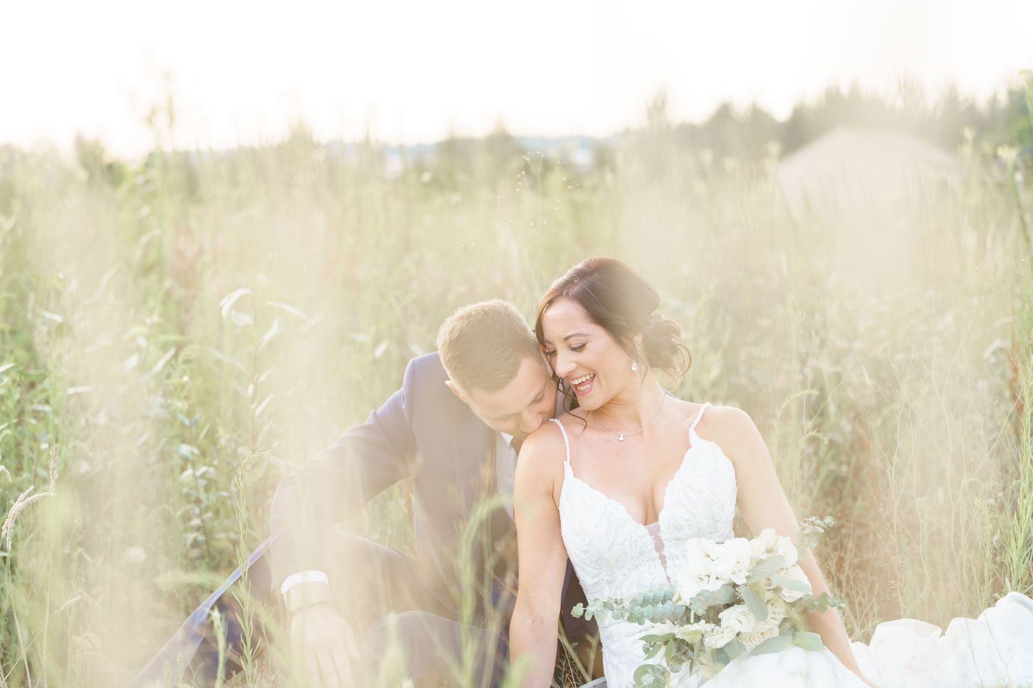 A romantic couple embraces in a dreamy outdoor setting with soft sunlight filtering through trees and tall grass.