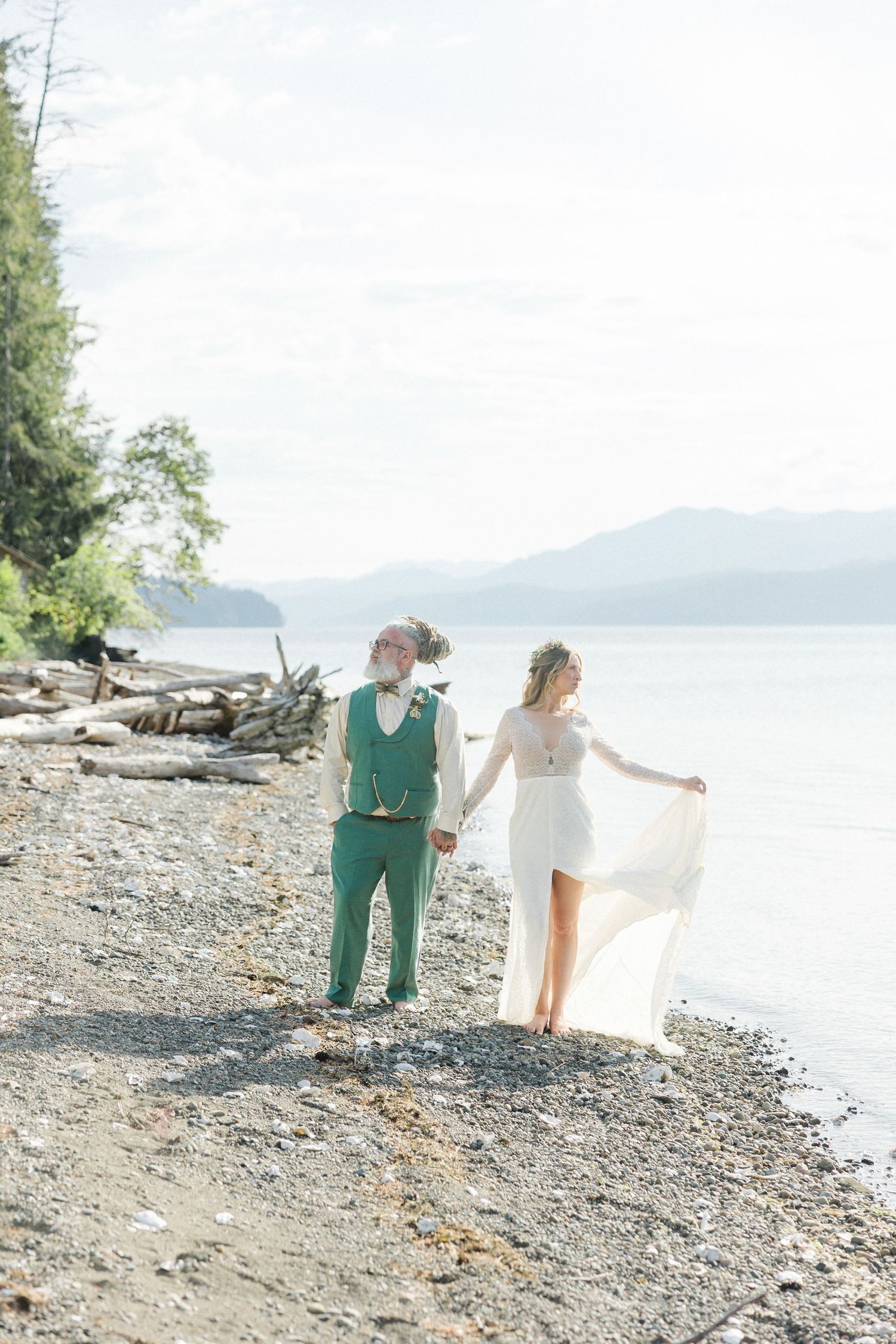 Bride and groom holding hands on rocky beach shoreline, mountains in background, bride in white dress, groom in teal suit.