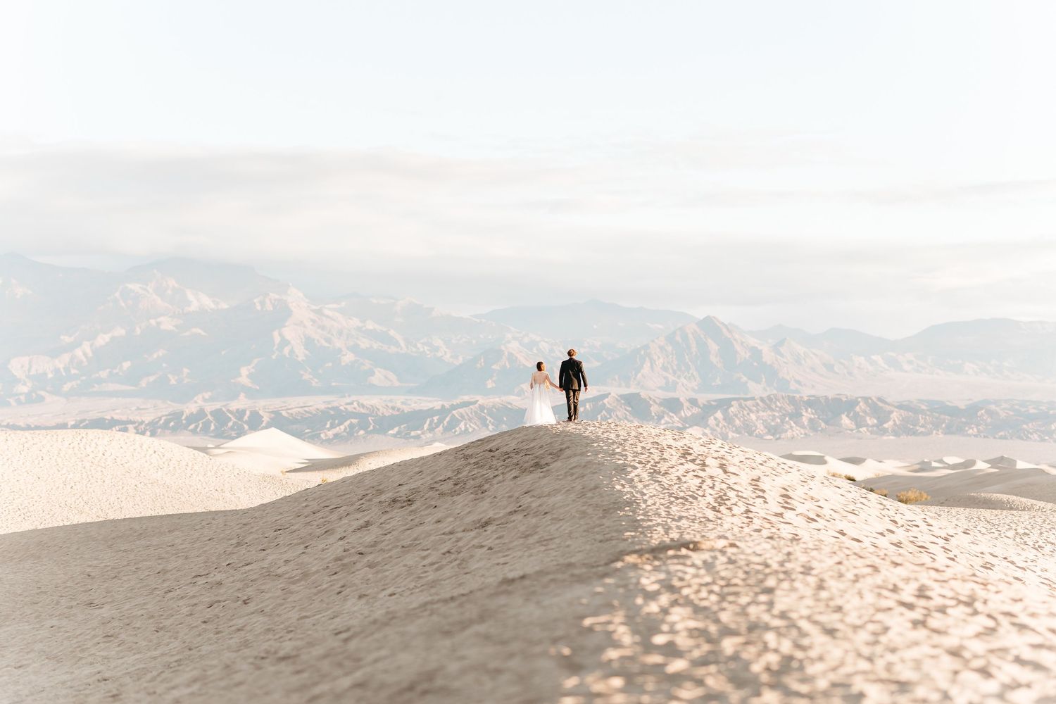 Two silhouettes walk along a sandy ridge with mountains in the distance under a soft, hazy sky.