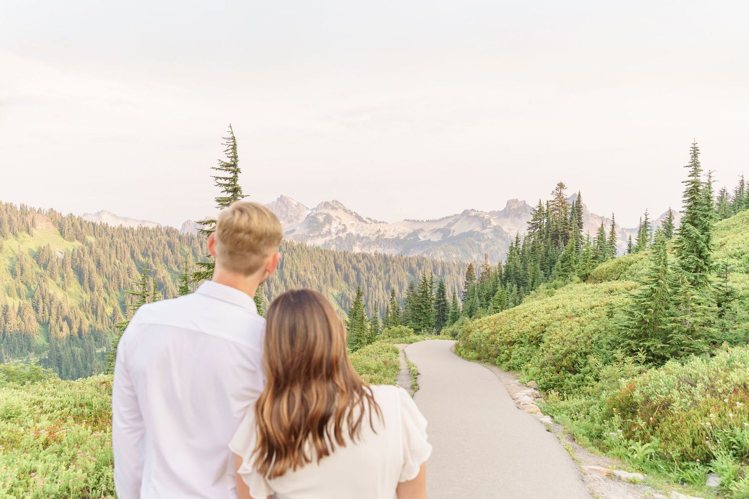 A couple stands on a scenic mountain trail overlooking evergreen forests and distant peaks at Mount Rainier National Park.