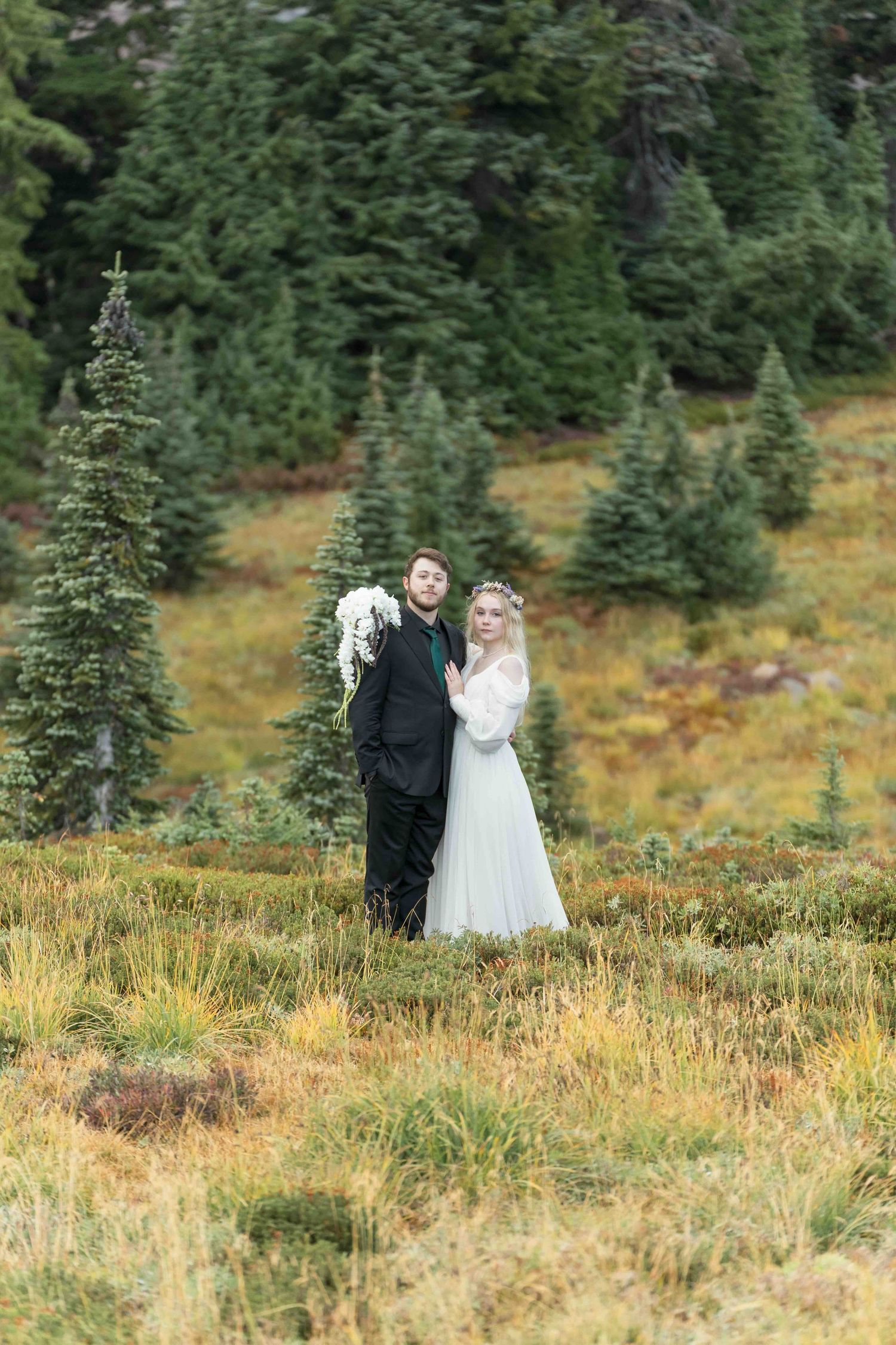 A couple in wedding attire stands in a mountain meadow surrounded by evergreen trees and autumn grasses.