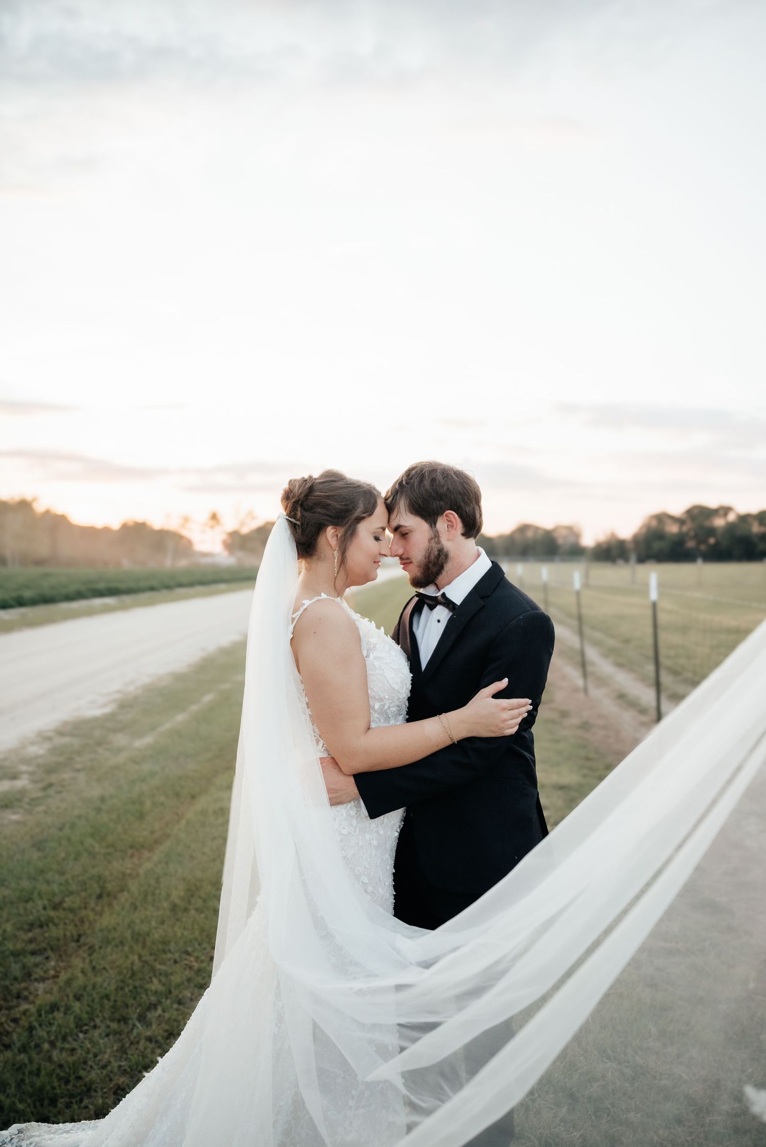 A romantic bride and groom touch foreheads at sunset on a rural road with veil flowing.