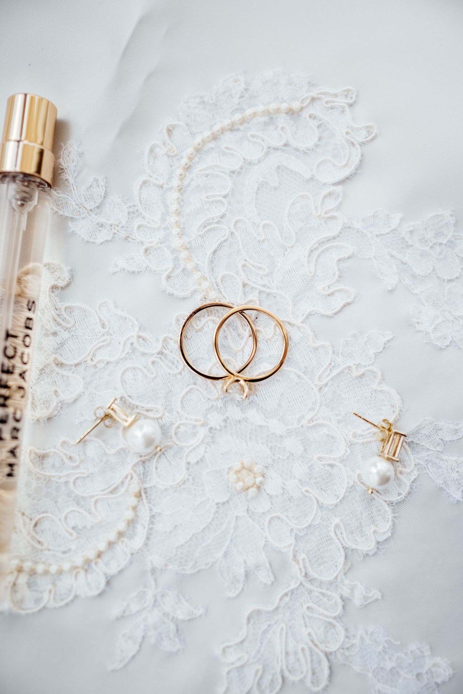 Gold wedding rings, pearl earrings, and Marc Jacobs perfume arranged on white bridal lace fabric.
