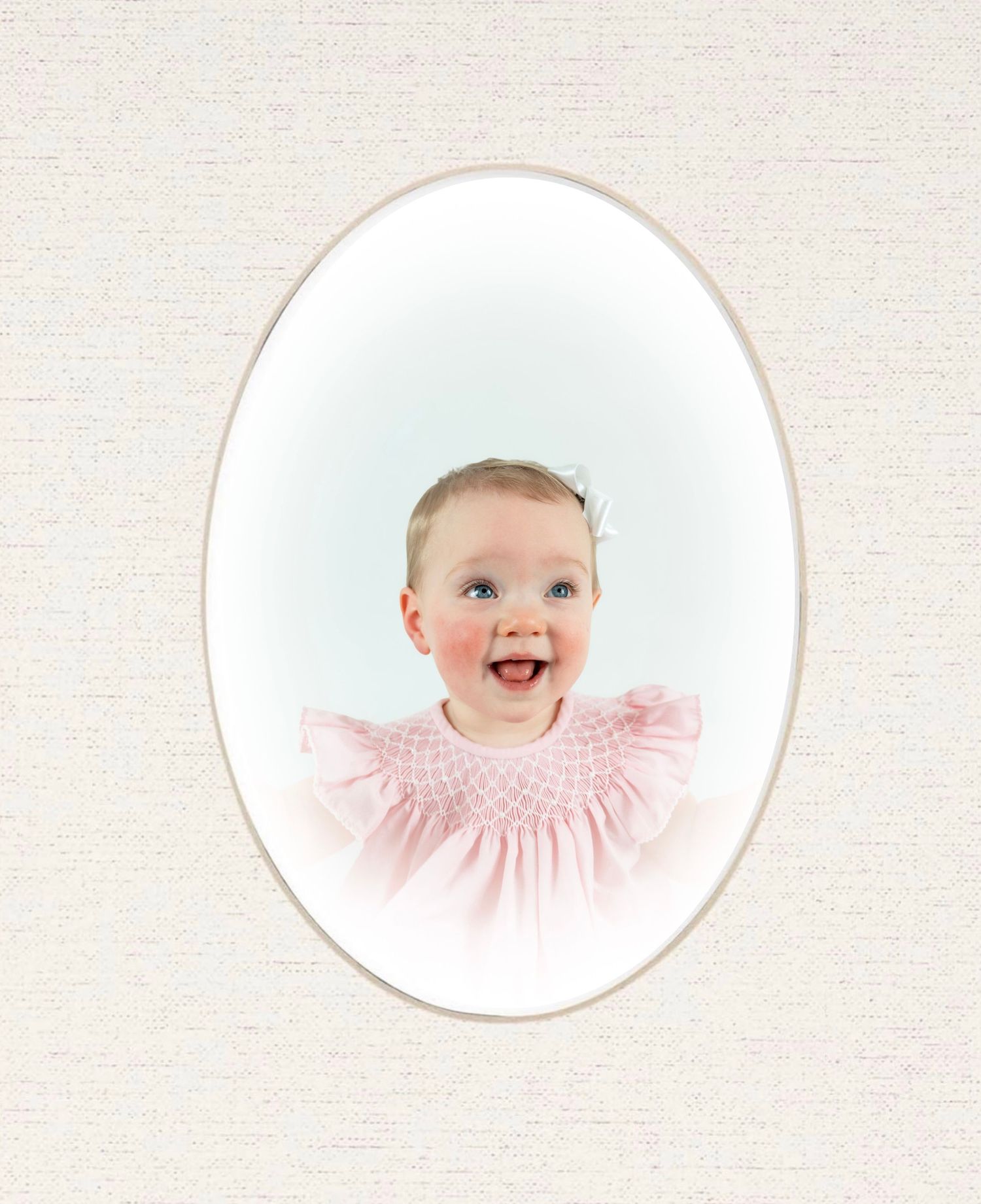 Smiling baby girl in pink smocked dress posed in classic oval portrait frame on cream textured background.