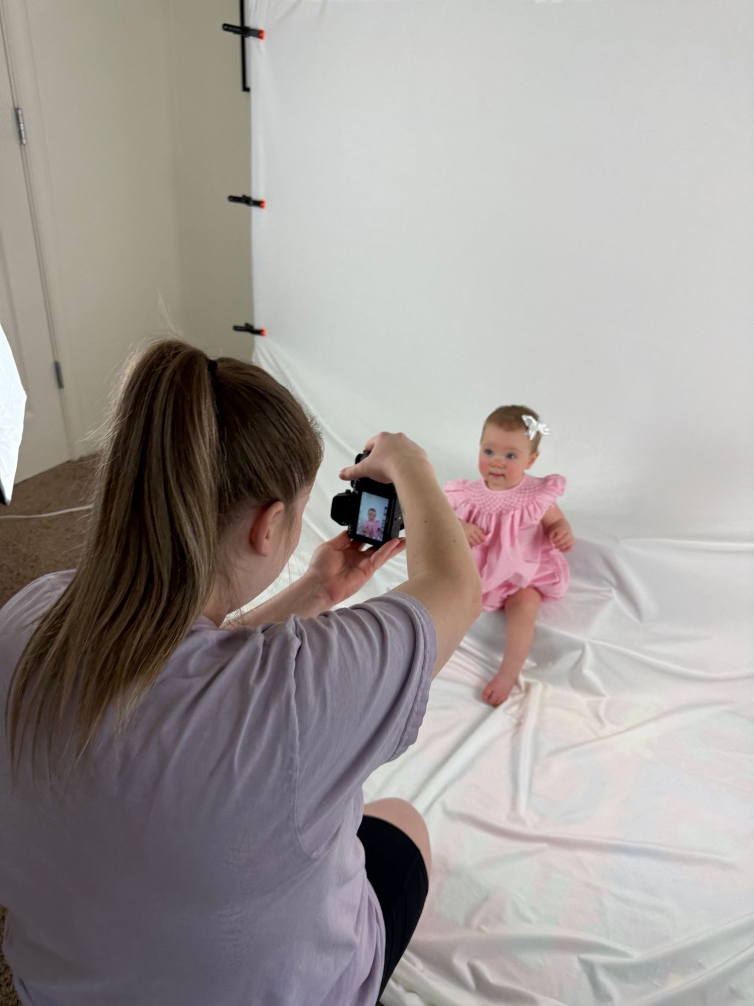Photographer taking pictures of a baby girl in a pink dress against a white backdrop in a home studio.