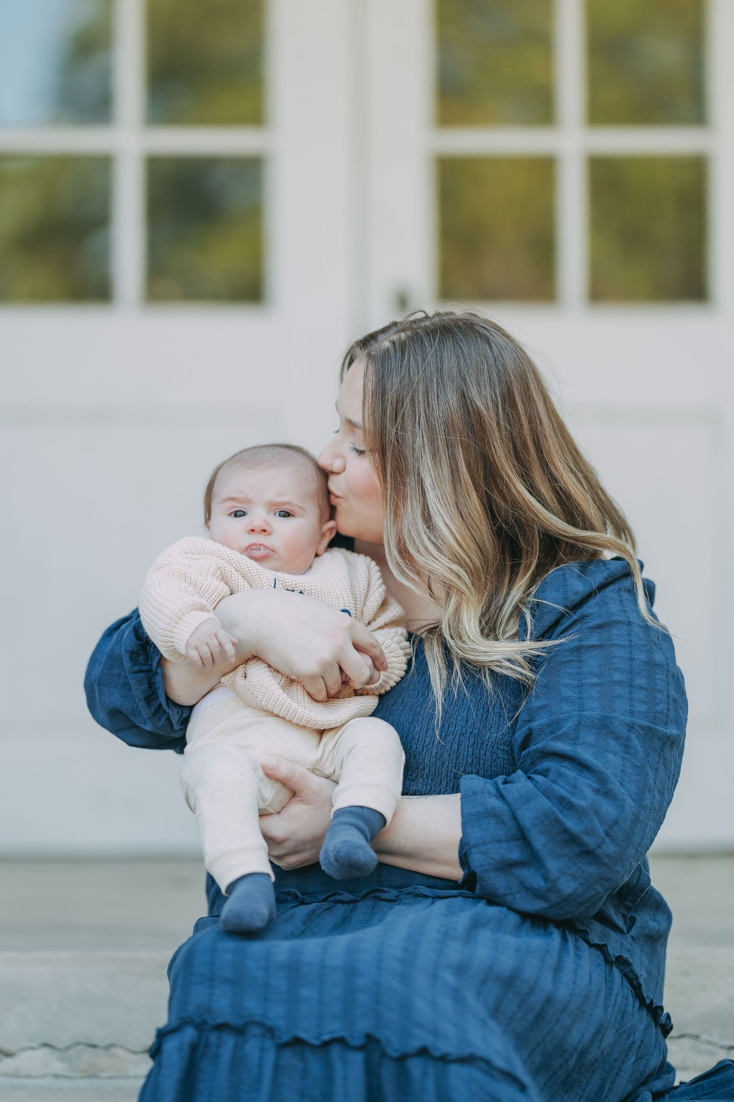 Mother in blue dress tenderly kissing her infant baby on the cheek while seated outdoors near white French doors.