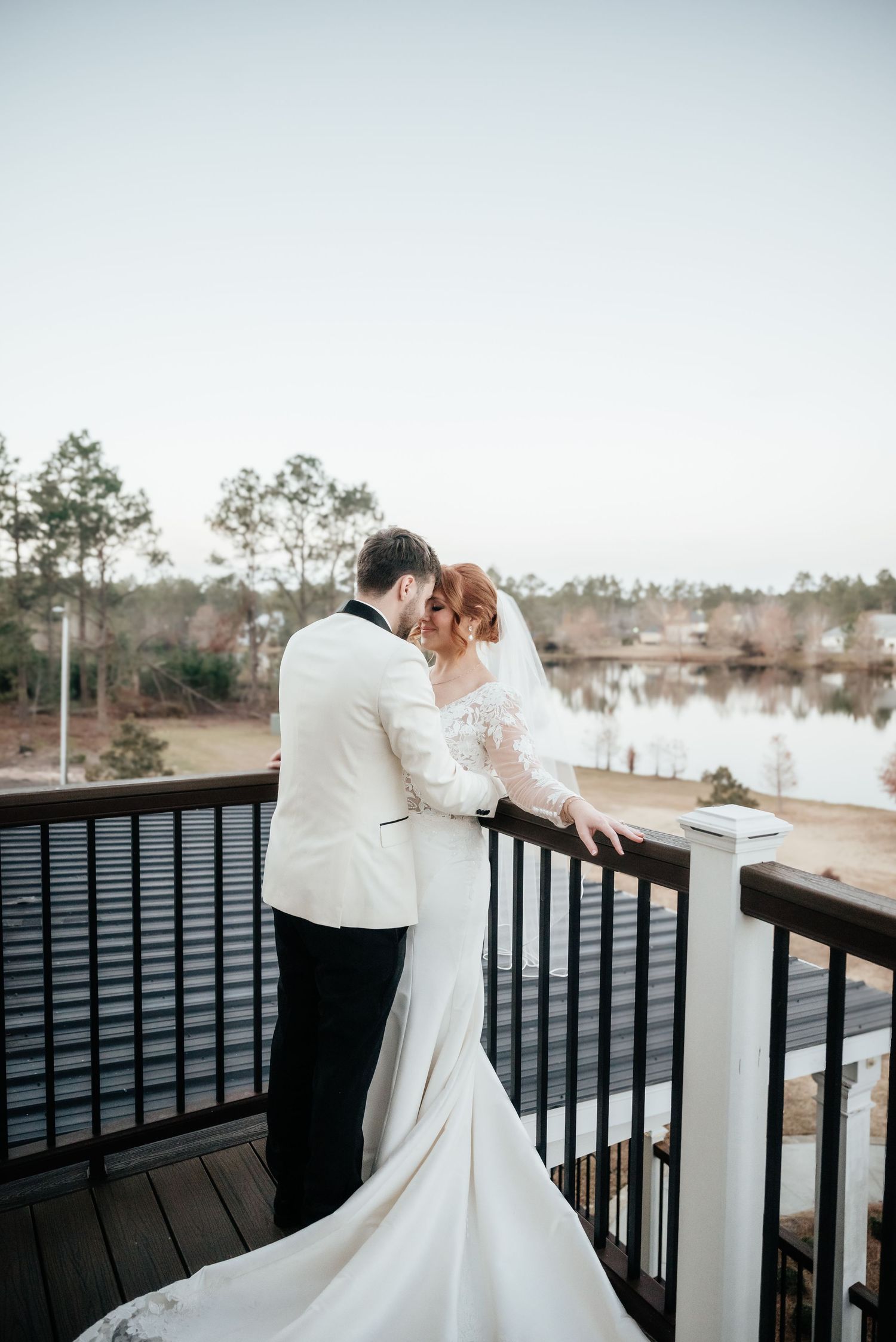 Bride and groom share a romantic embrace on a deck overlooking a serene lake at sunset.