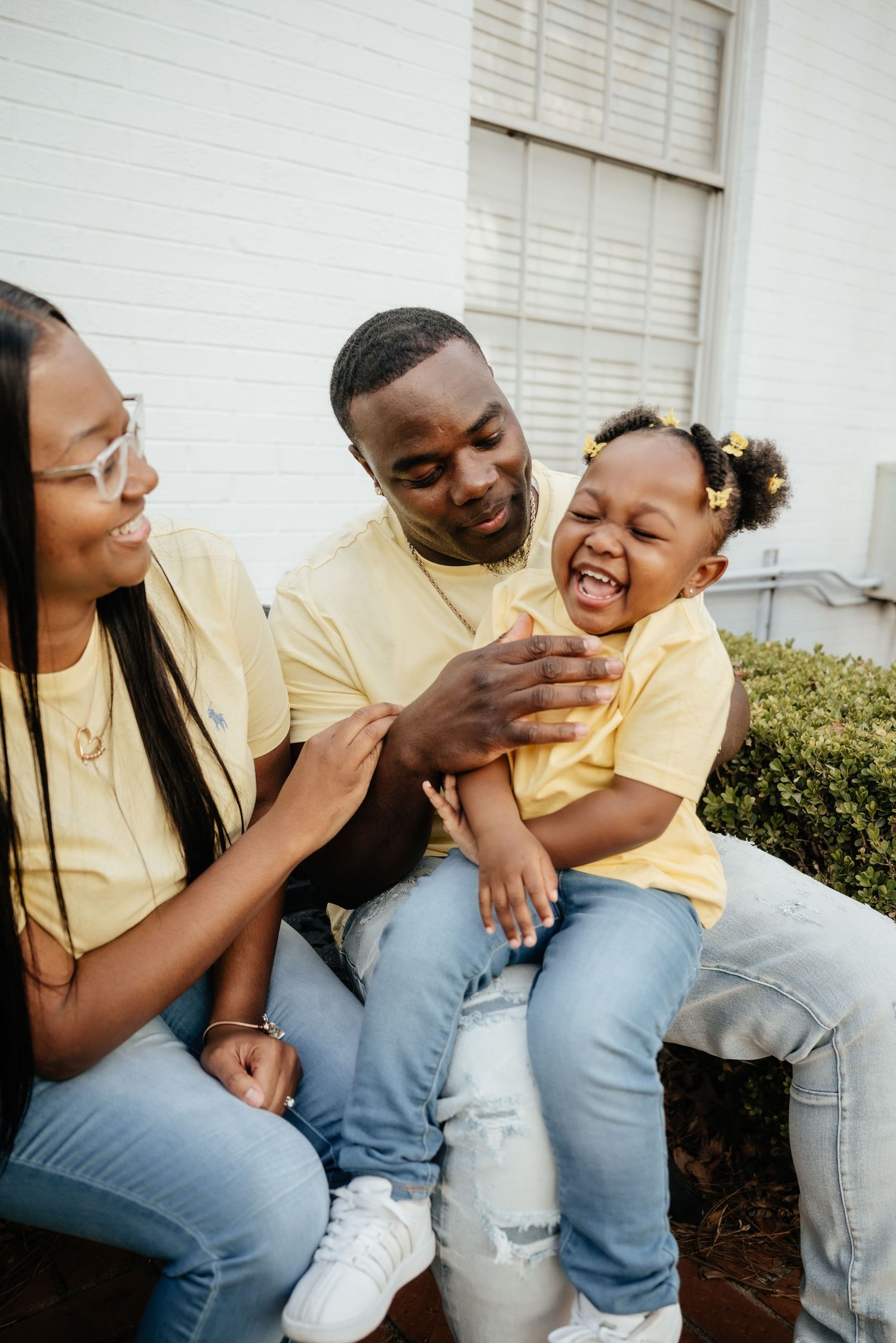 Joyful Black family in matching yellow shirts and jeans laughing together outdoors near a white brick building.