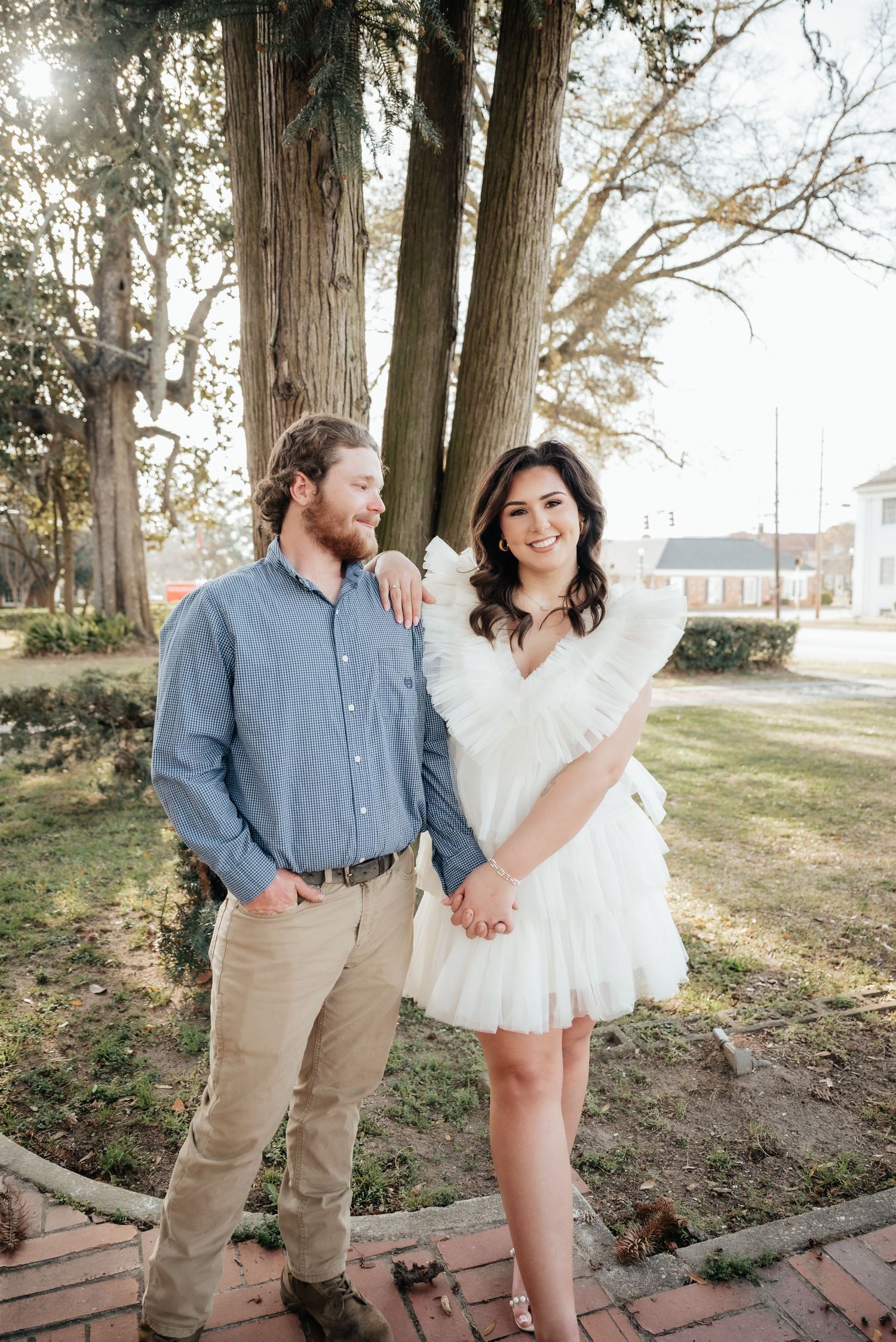 A couple holds hands and smiles while standing near large trees on a brick sidewalk outdoors.