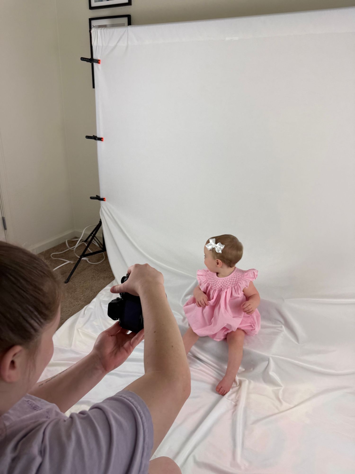 Photographer taking pictures of a baby girl in a pink dress sitting on a white backdrop in a home photography studio.