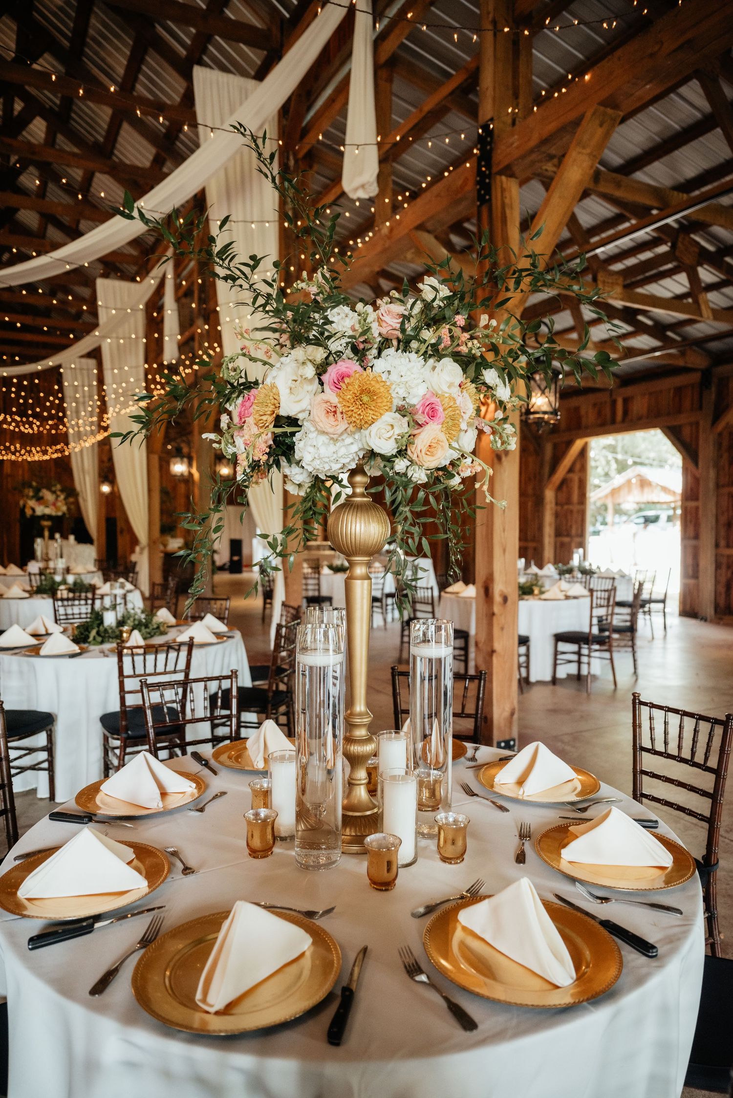 Elegant barn wedding reception with gold chargers, white napkins, tall floral centerpieces, string lights, and draped fabric decor.