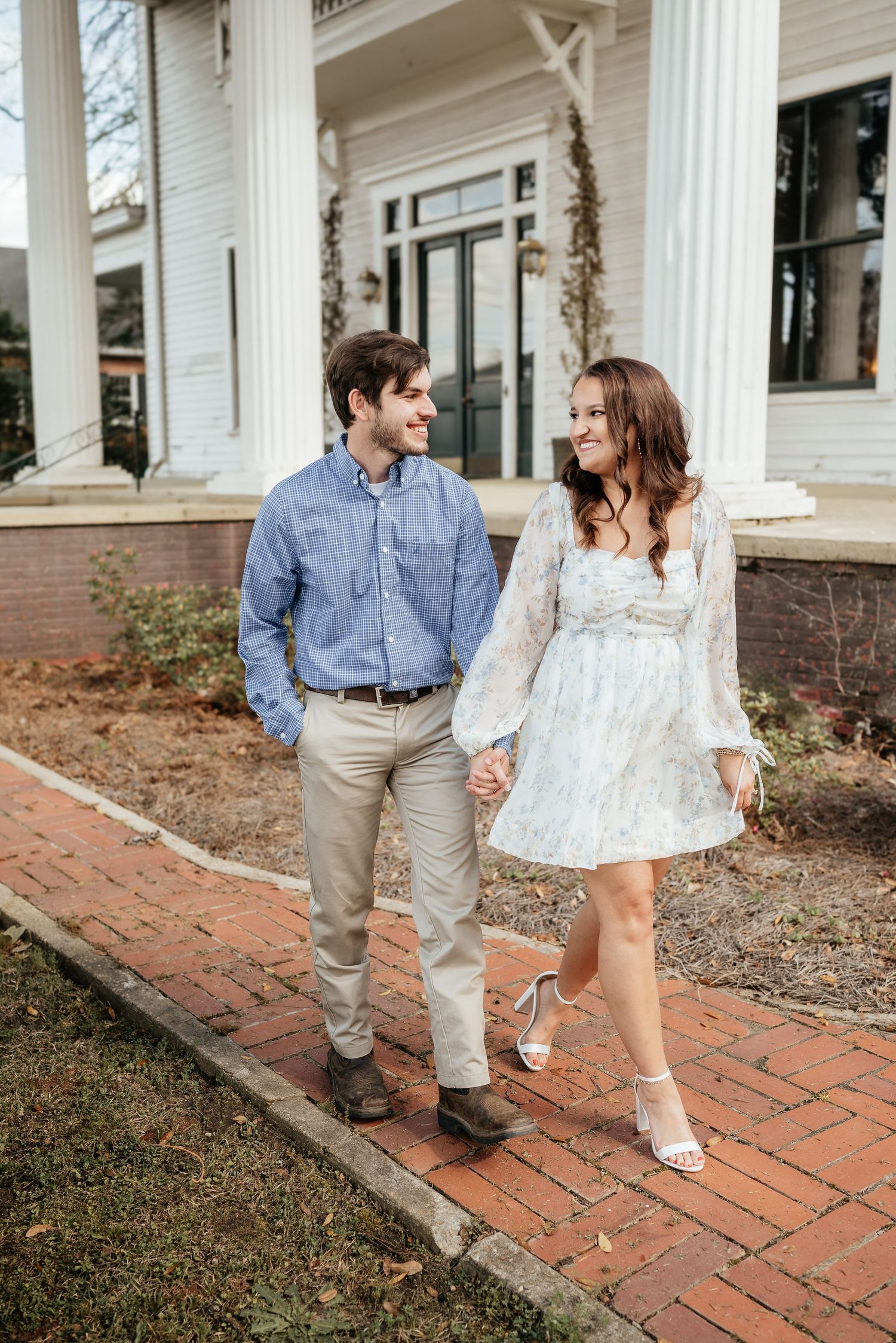A smiling couple walks hand-in-hand along a brick path in front of a white colonial building.