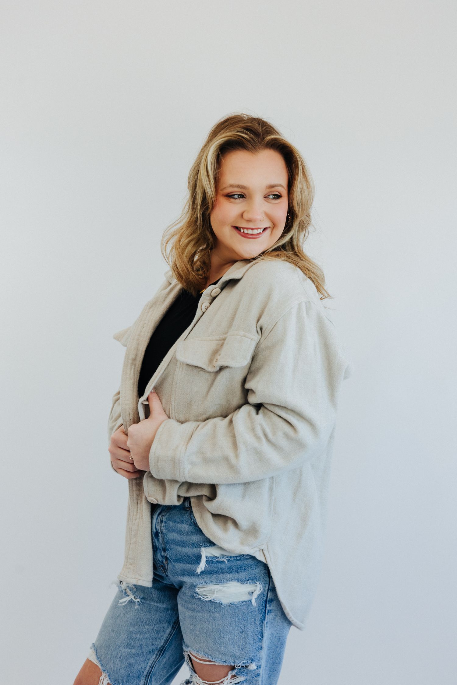 Happy young woman in distressed denim and beige shacket poses sideways in a bright studio.