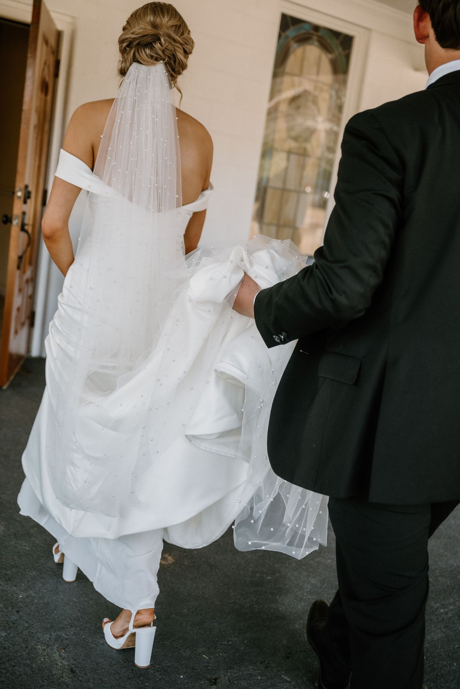 A bride in a white off-shoulder gown with veil walks with groom in dark suit entering a venue.