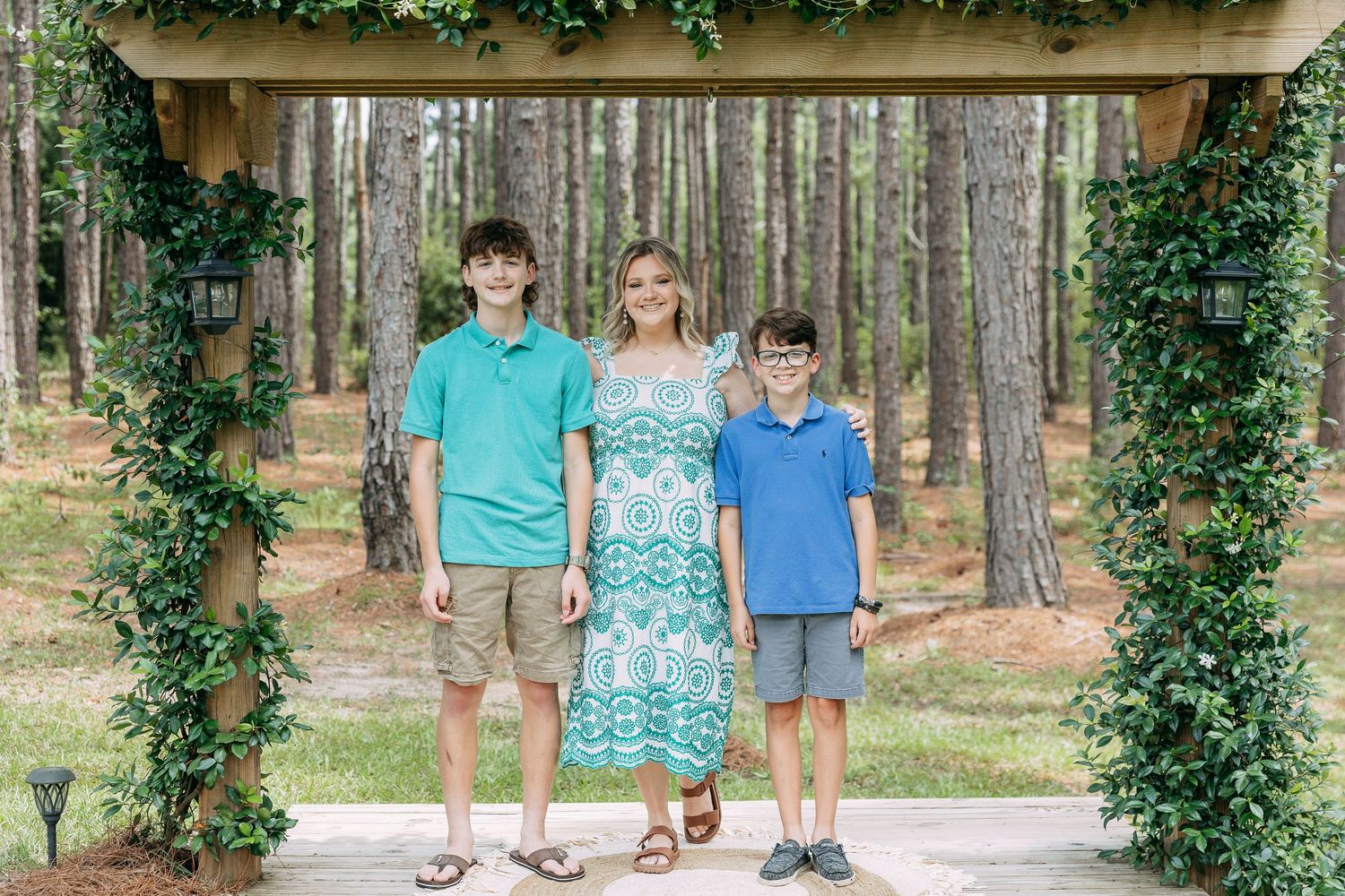 A woman and two boys smile together under a lush ivy-covered pergola in a pine forest.