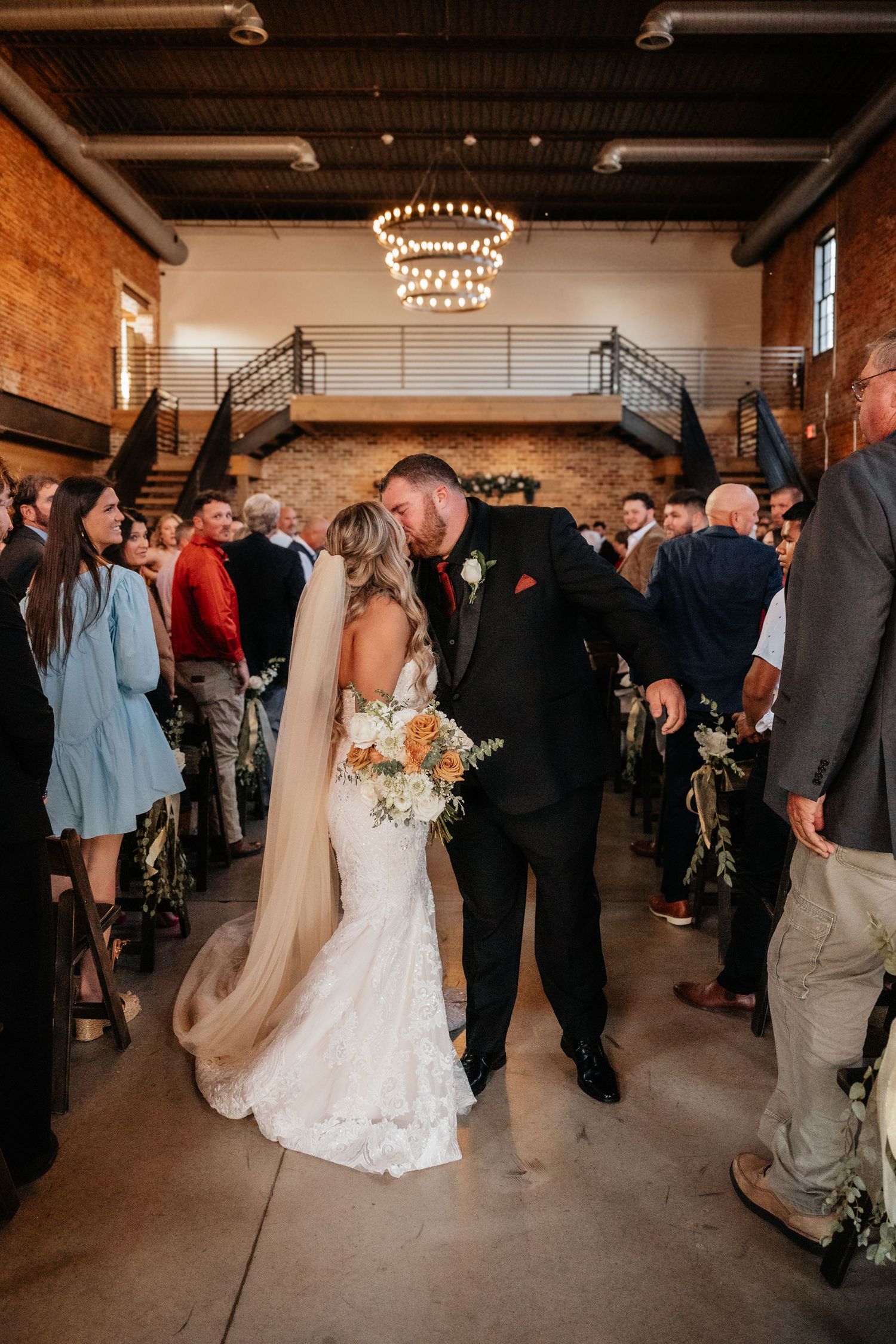 A newlywed couple shares a kiss while walking down the aisle in a rustic industrial wedding venue.