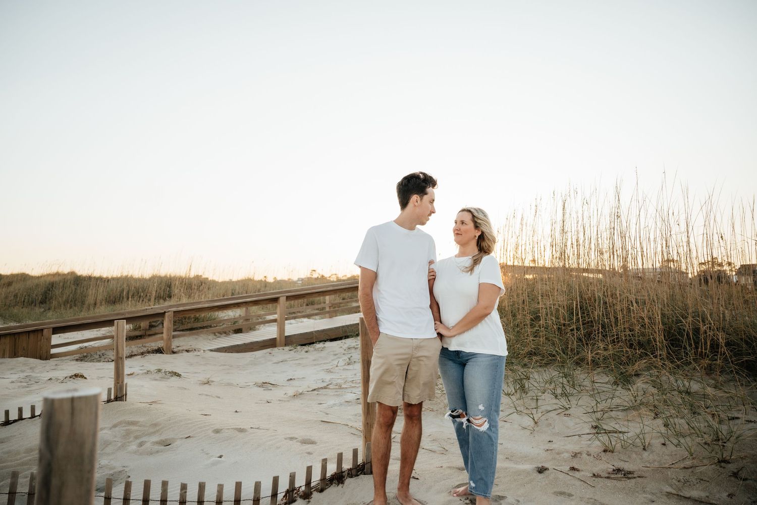 A happy couple holds hands on a sandy beach boardwalk at golden hour sunset.