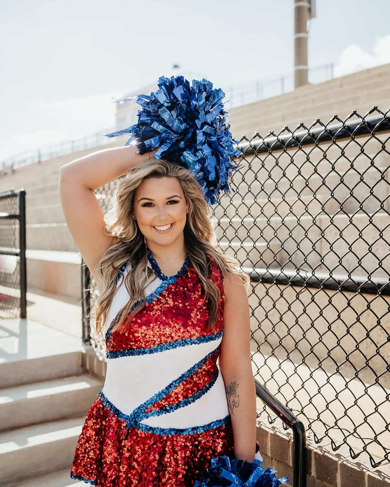 Smiling cheerleader in red sequin uniform holding blue pom-poms at a stadium bleacher backdrop.