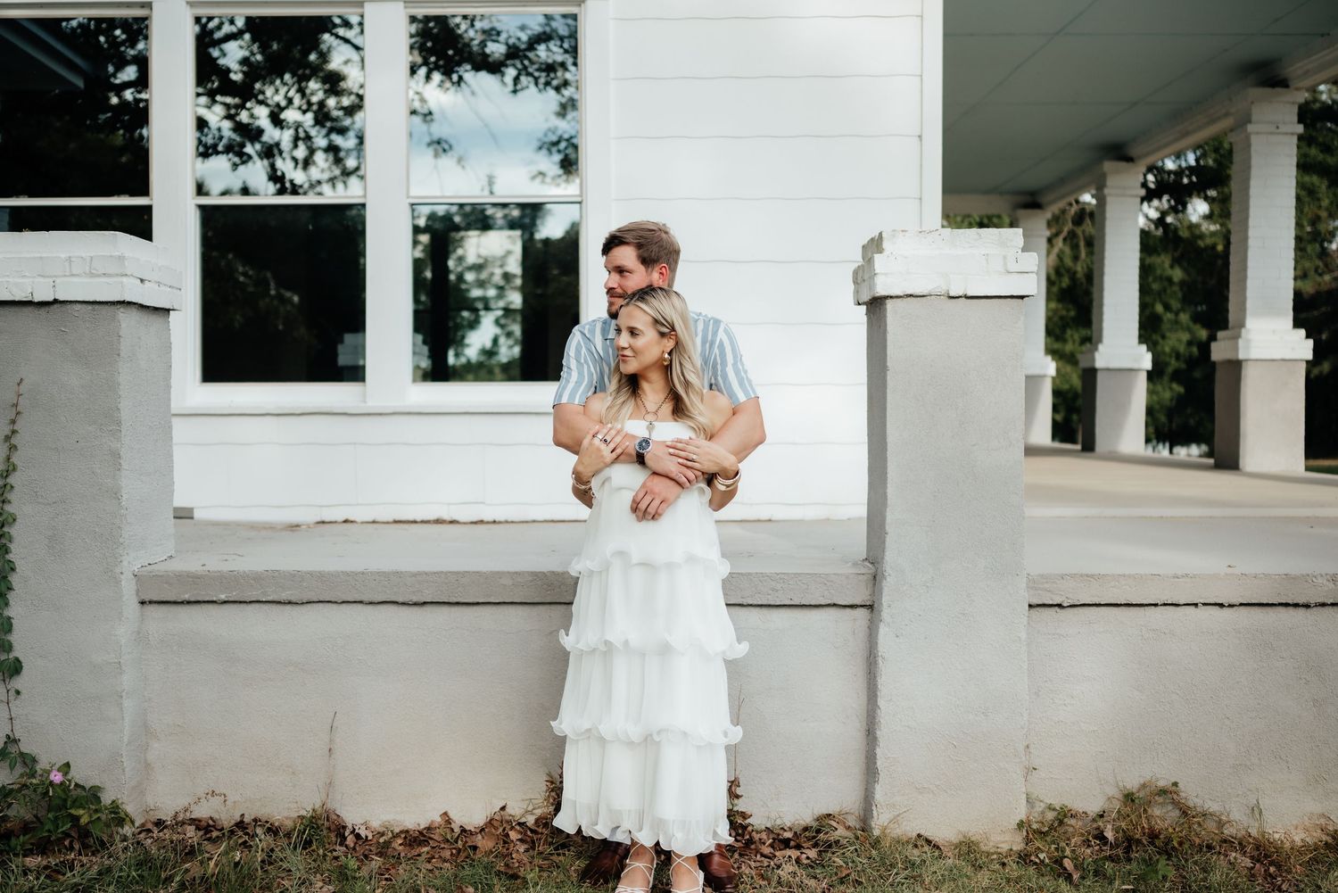 Couple embracing in front of a white farmhouse porch, woman in white tiered dress, man in striped shirt outdoors.