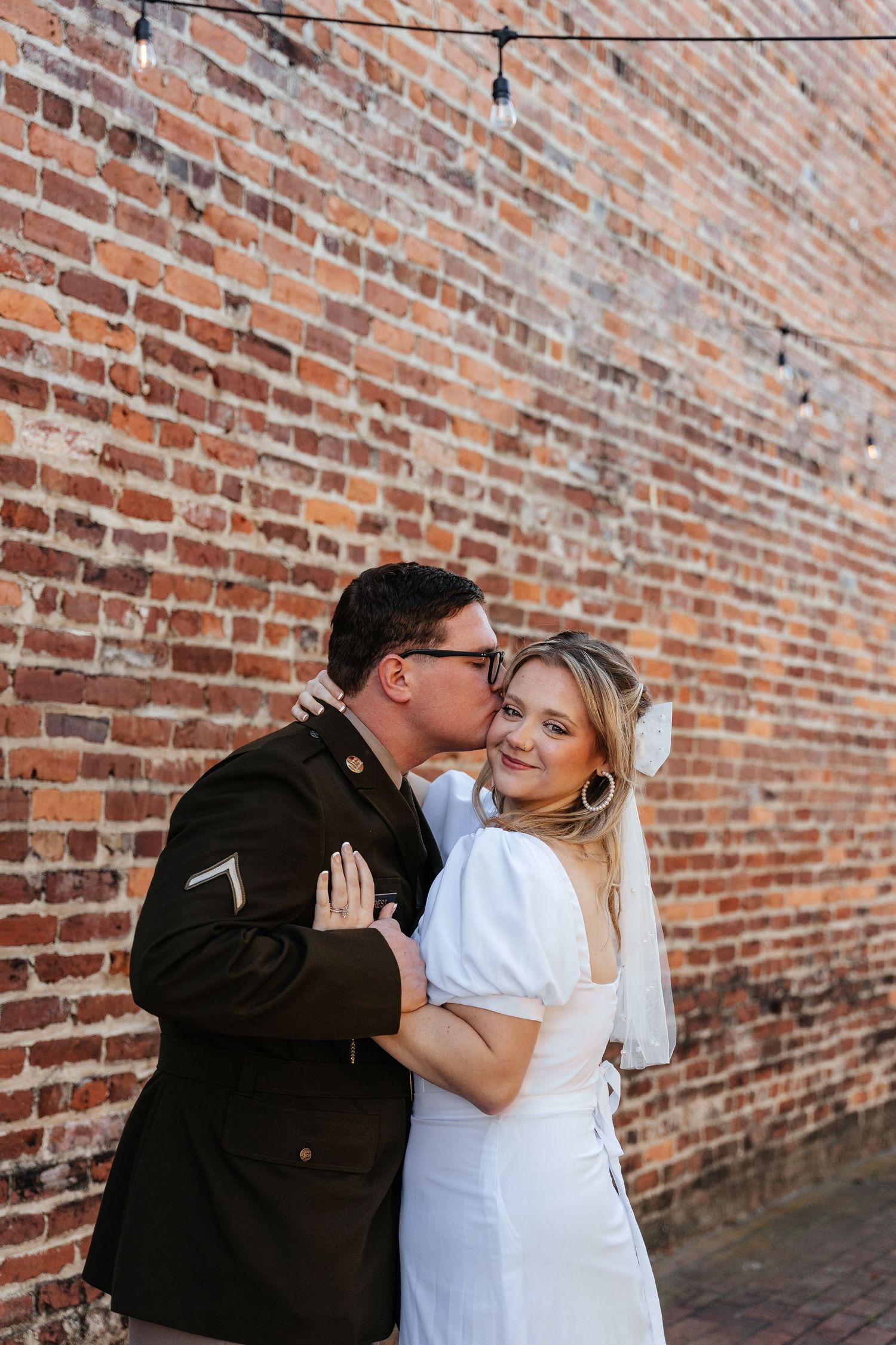 Bride and groom share a romantic kiss against a rustic brick wall with string lights at their wedding.