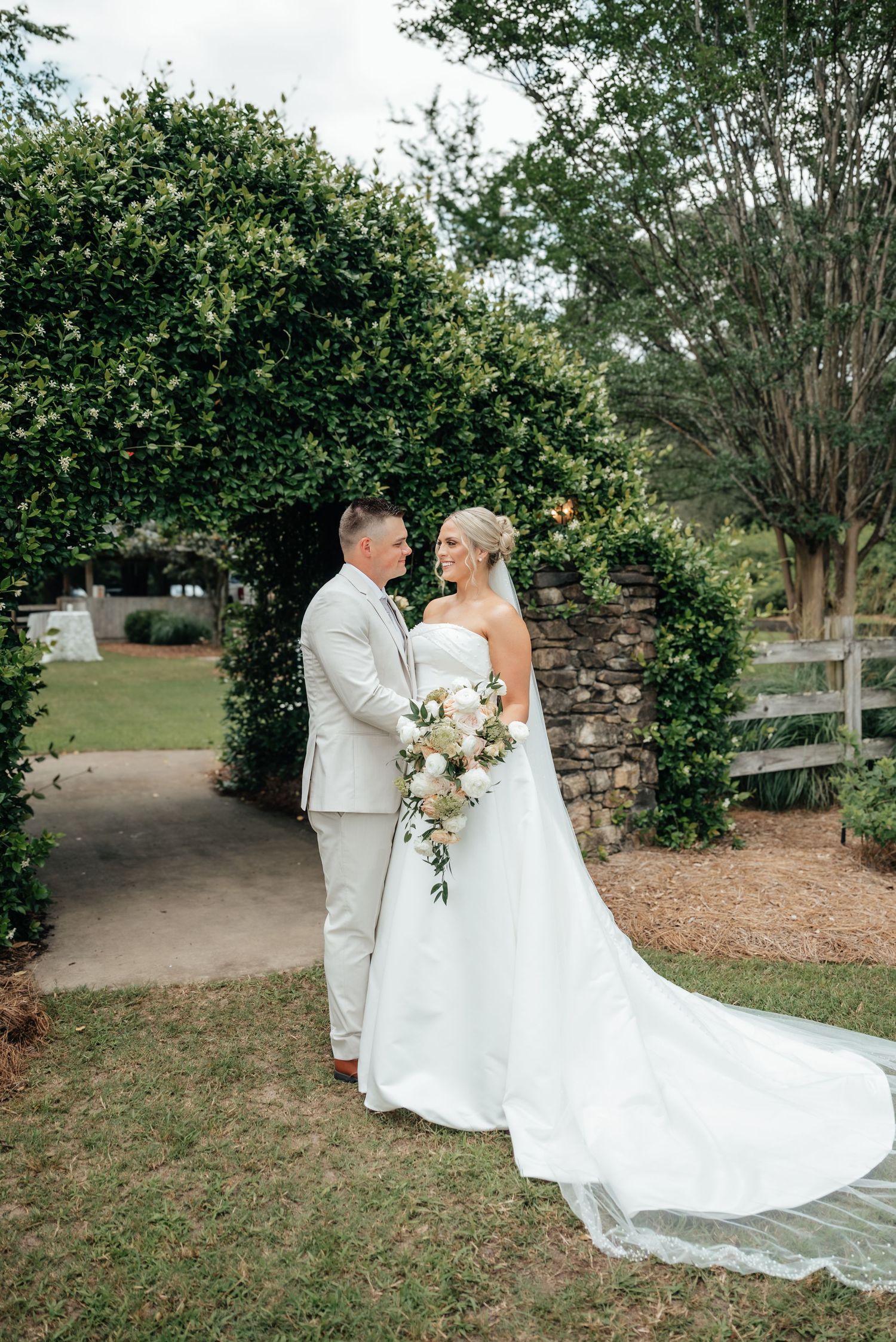Bride and groom stand together under a lush green archway holding a cascading wedding bouquet.