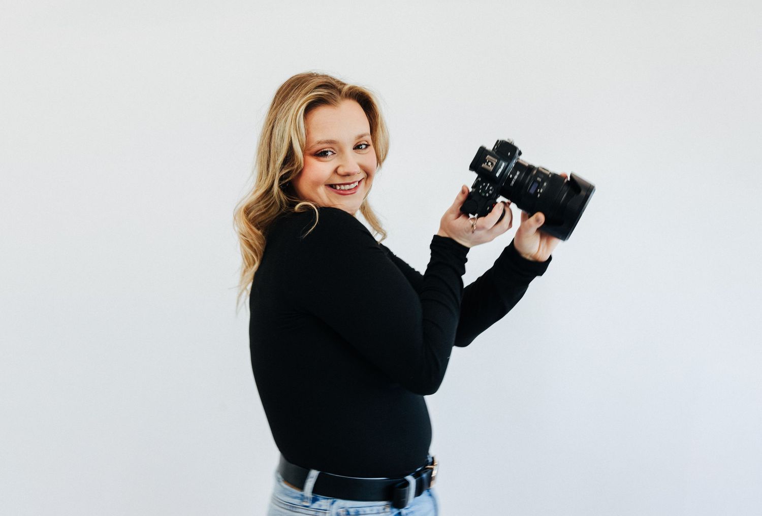 Professional female photographer smiles while holding a camera against a clean white studio background.