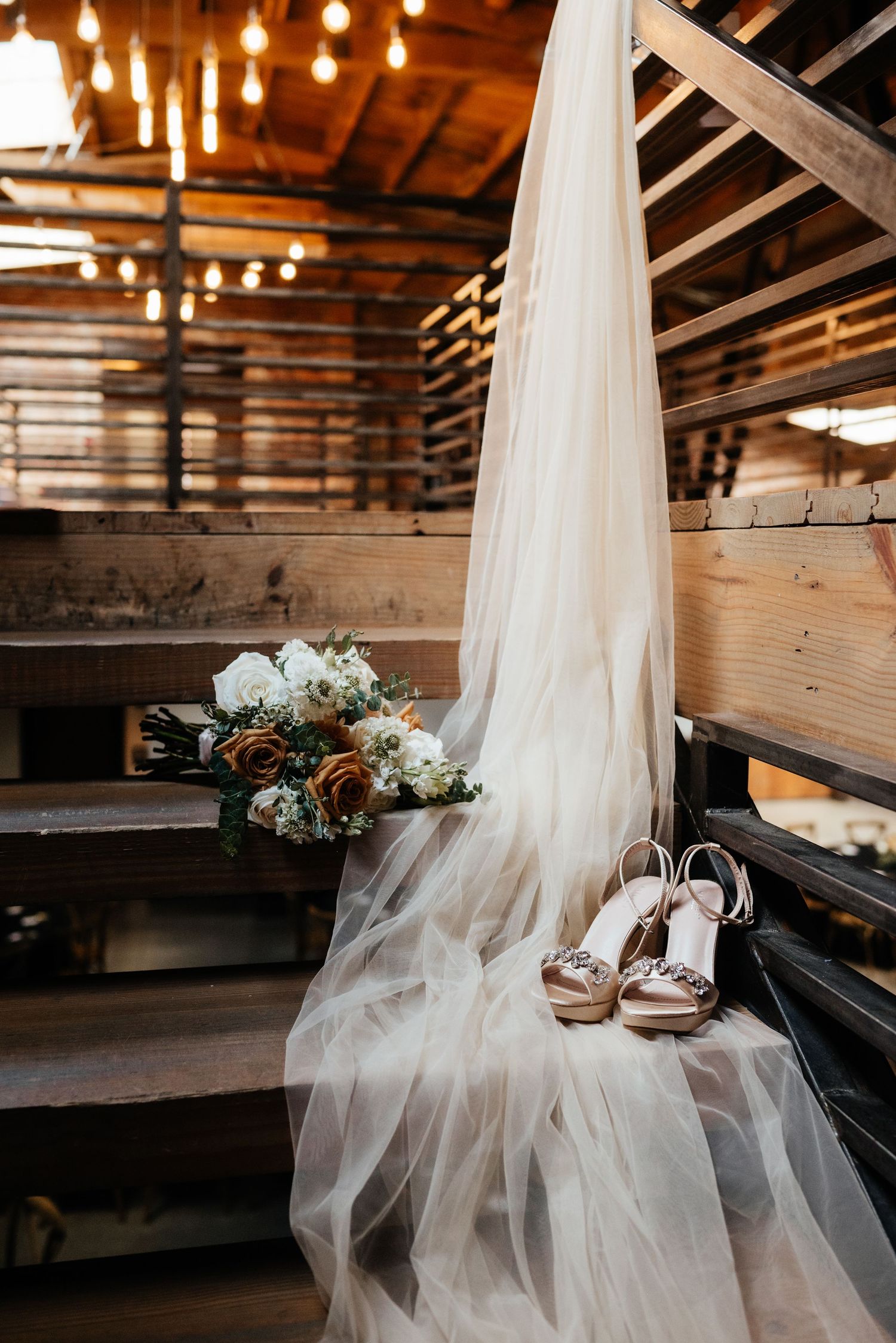 Elegant bridal veil, floral bouquet, and strappy heels arranged on wooden barn steps with string lights.
