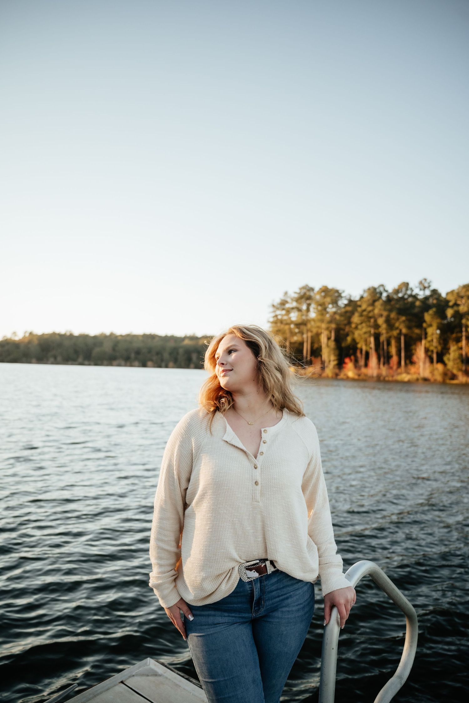 Young blonde woman in cream henley and jeans standing on a lake dock at golden hour sunset.