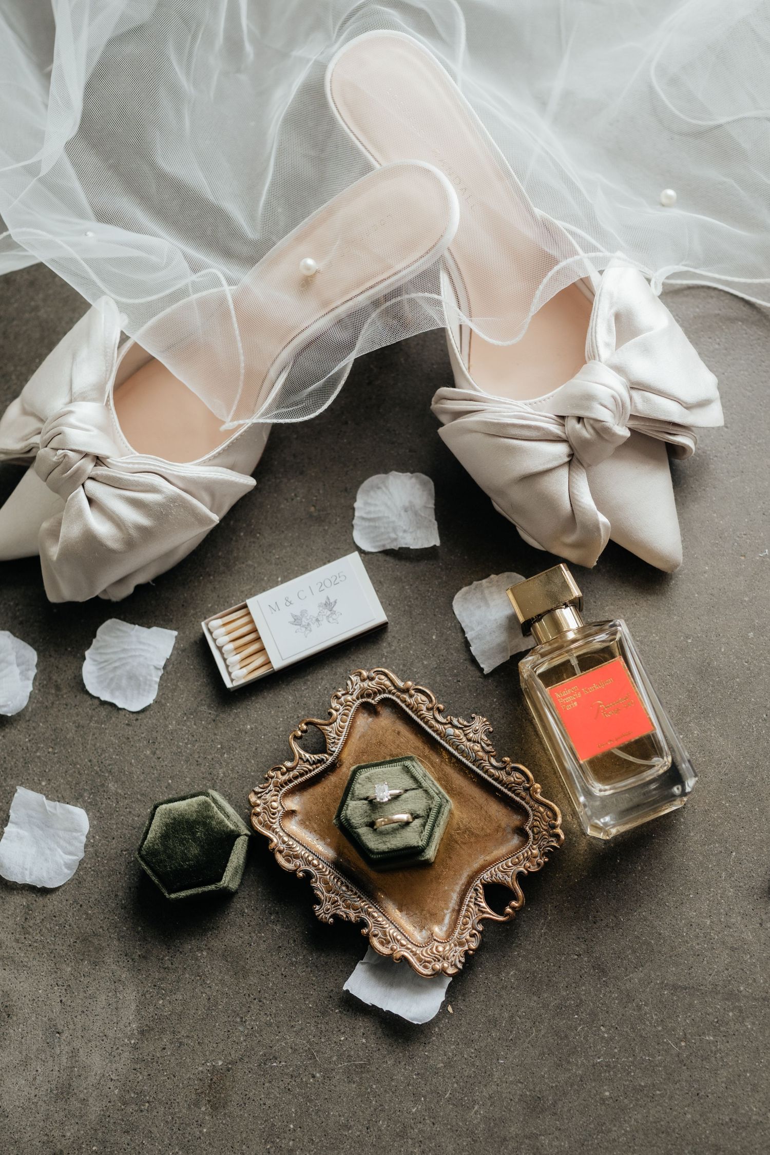 Bridal flat lay with white bow heels, engagement ring in velvet box, perfume, and rose petals.