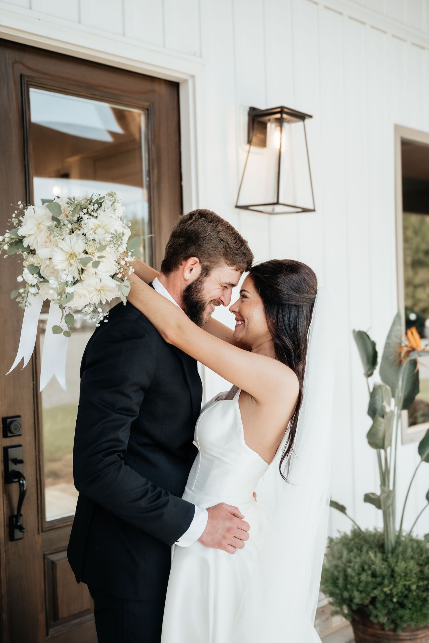 Bride and groom sharing an intimate moment outside a white farmhouse wedding venue with floral bouquet.