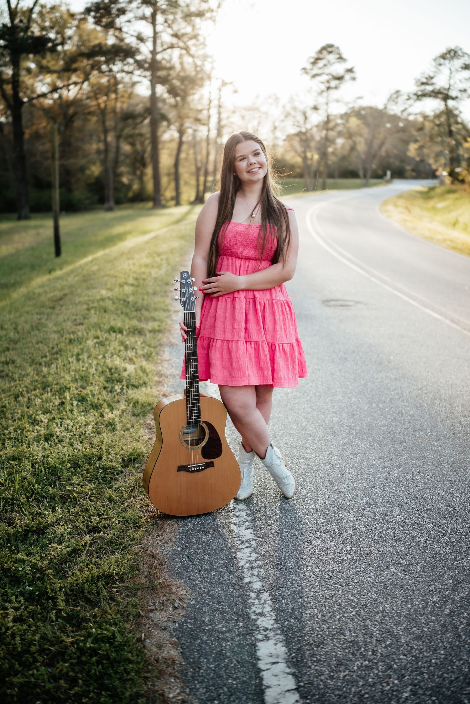 A smiling teenage girl in a pink dress holds an acoustic guitar while standing on a country road.
