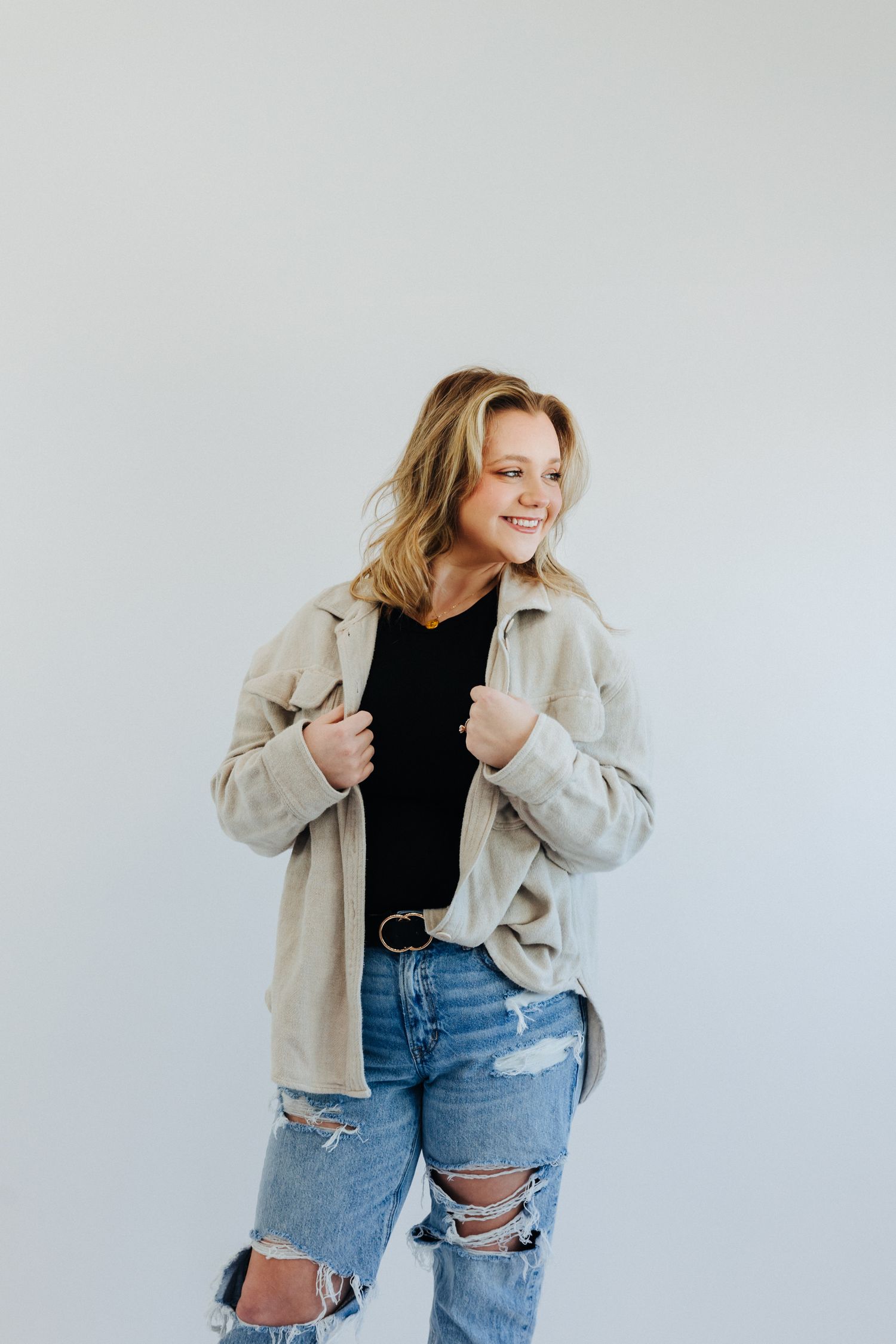 Smiling young woman in ripped jeans and beige jacket poses against a white studio background.