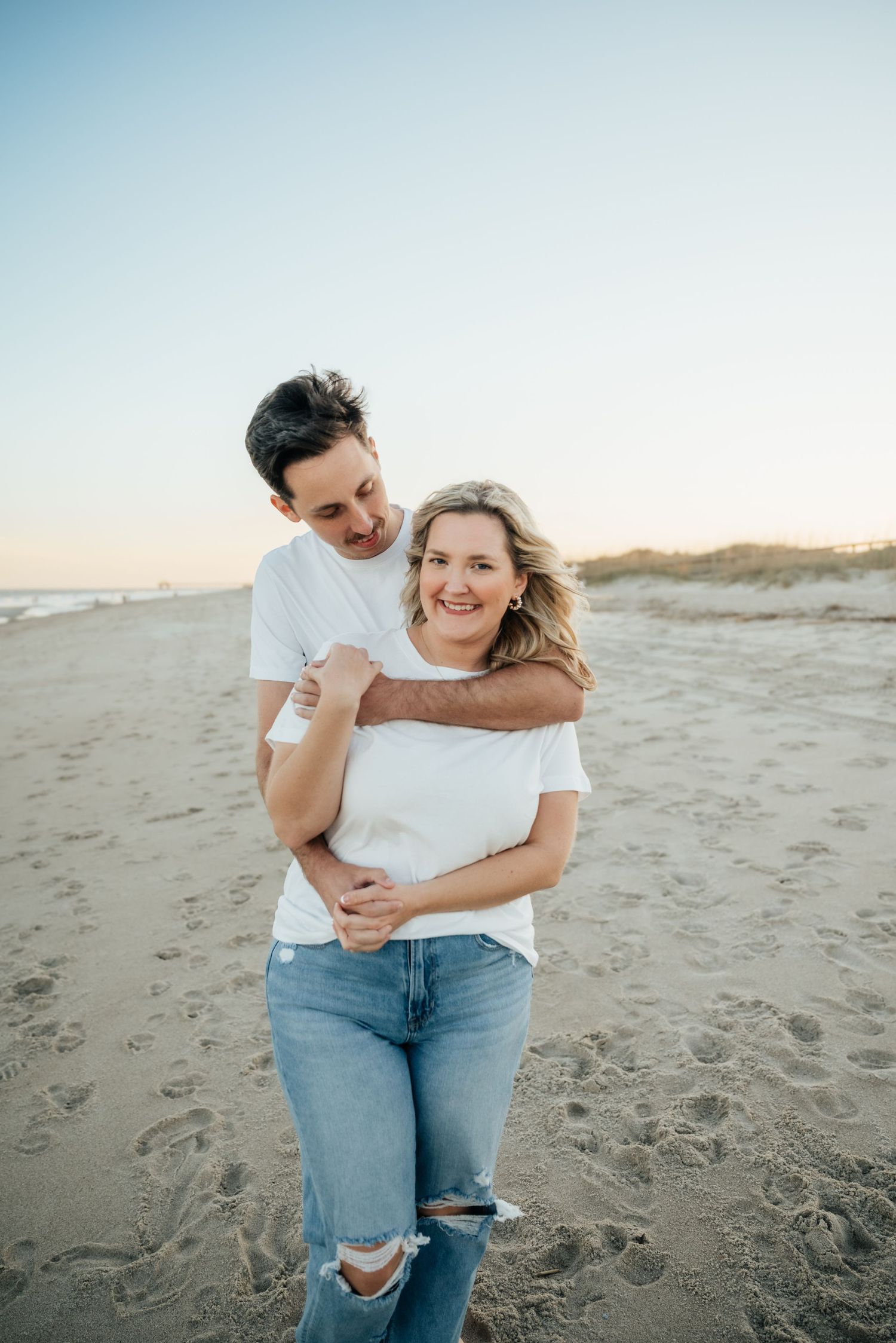 Happy couple embraces on a sandy beach at sunset wearing matching white tops and blue jeans.