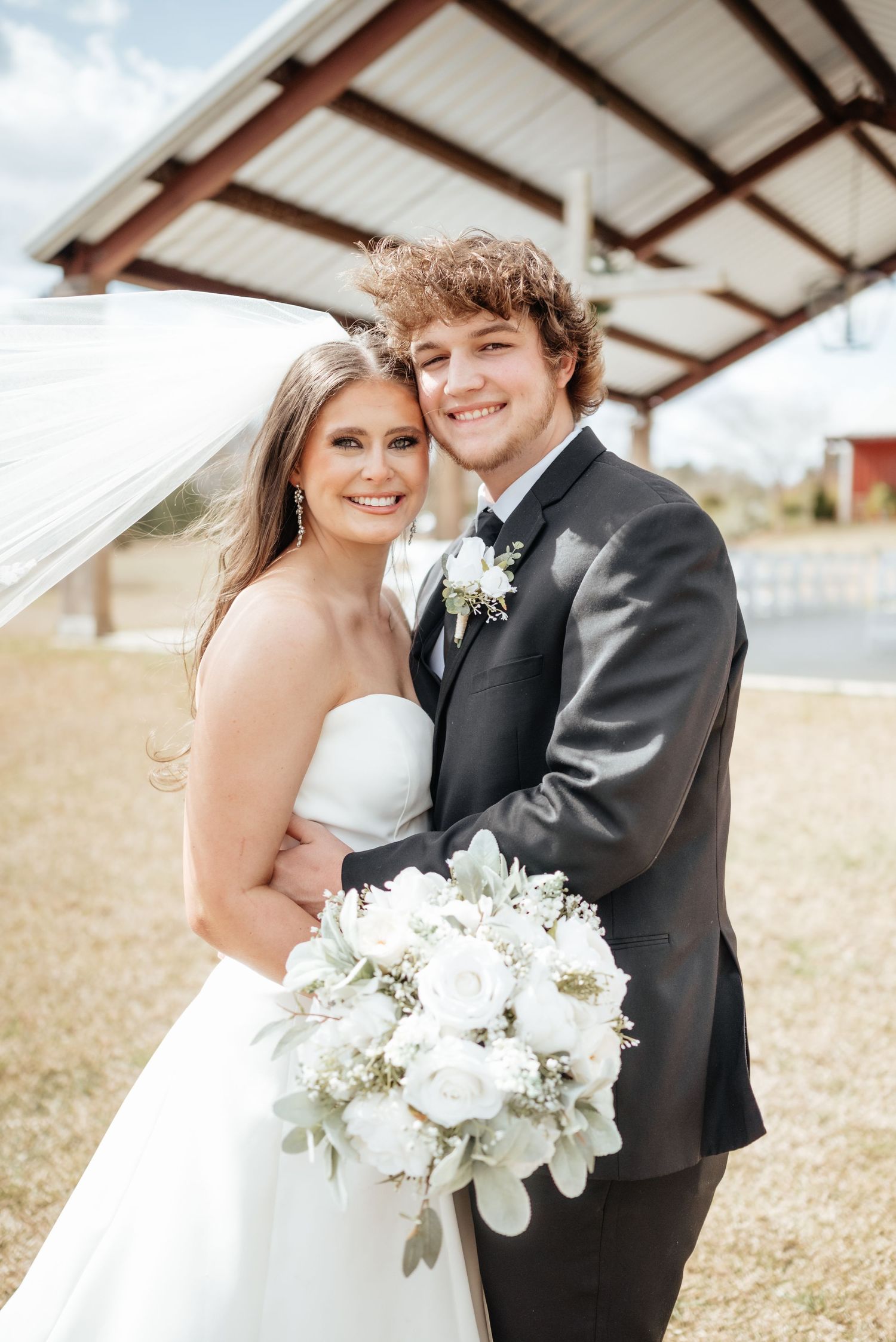 Newlyweds smiling outdoors, bride in strapless gown holding white bouquet, groom in charcoal suit.