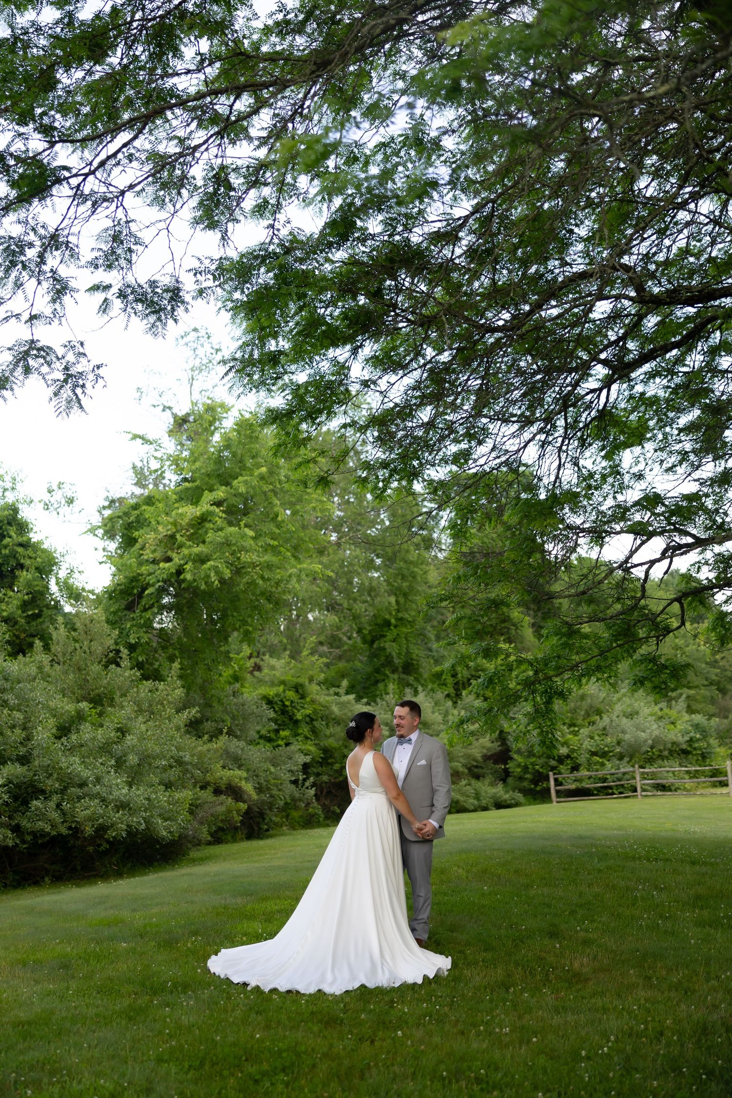 Bride and groom stand facing each other on lush green lawn beneath large tree canopy at outdoor wedding venue.