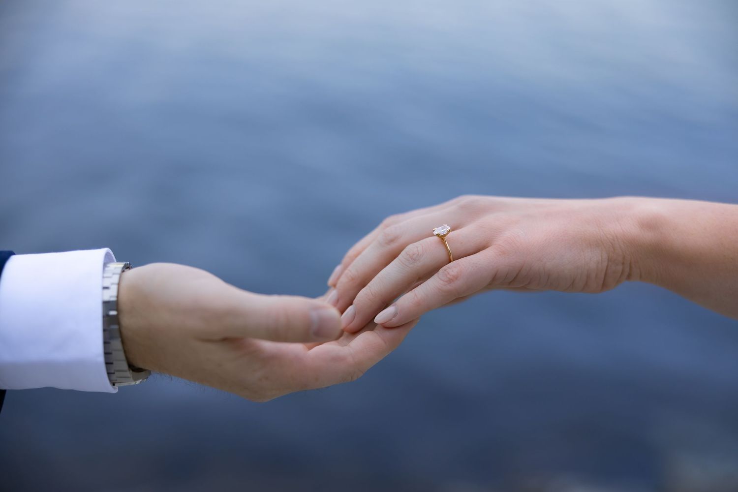 Couple holding hands outdoors near water, woman wearing a gold engagement ring, romantic proposal moment.