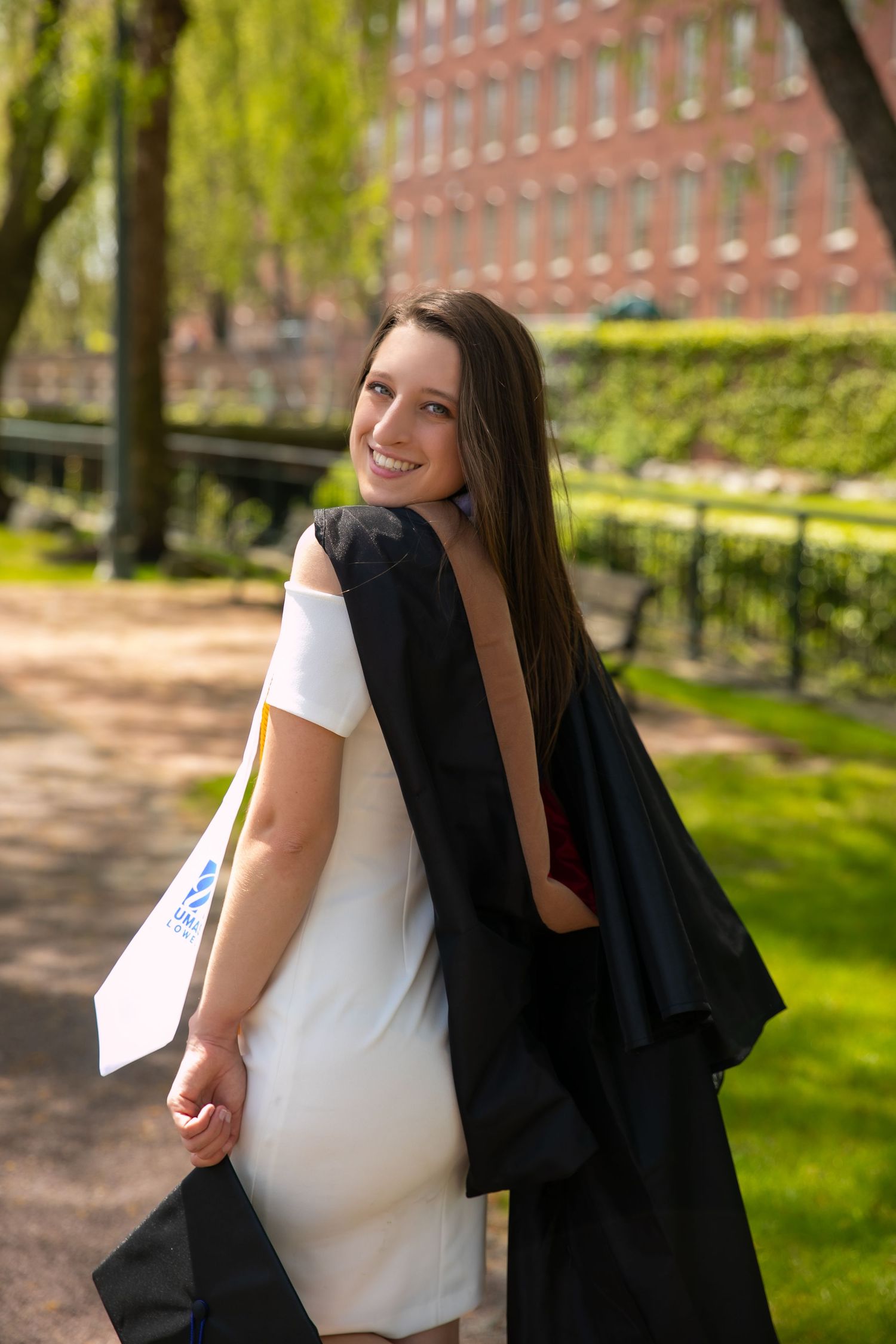 Graduate in white shirt and black gown poses outdoors on sunny spring day in university garden setting.