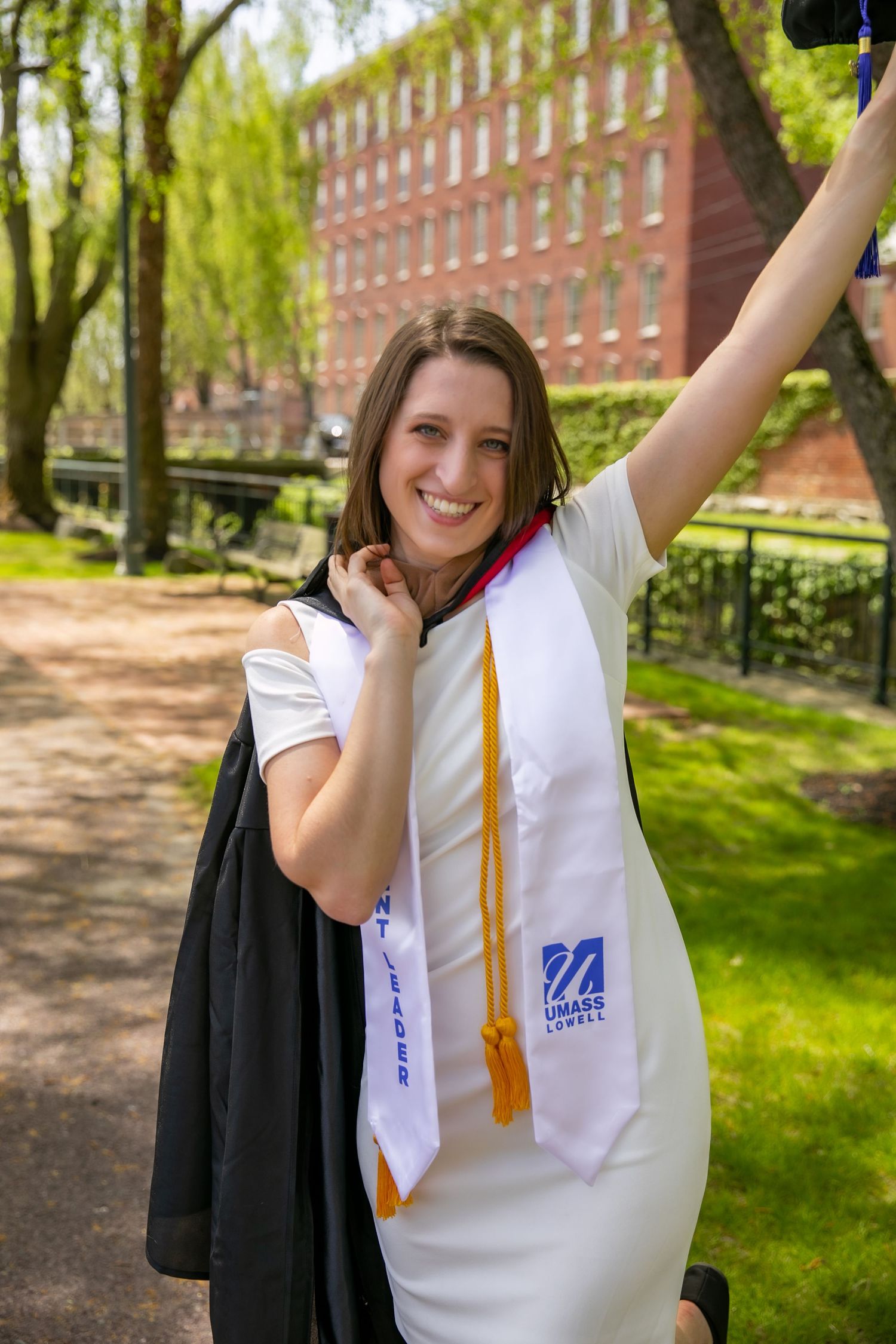 A graduate in white regalia celebrates with raised arms on a sunny spring day outside a brick university building.